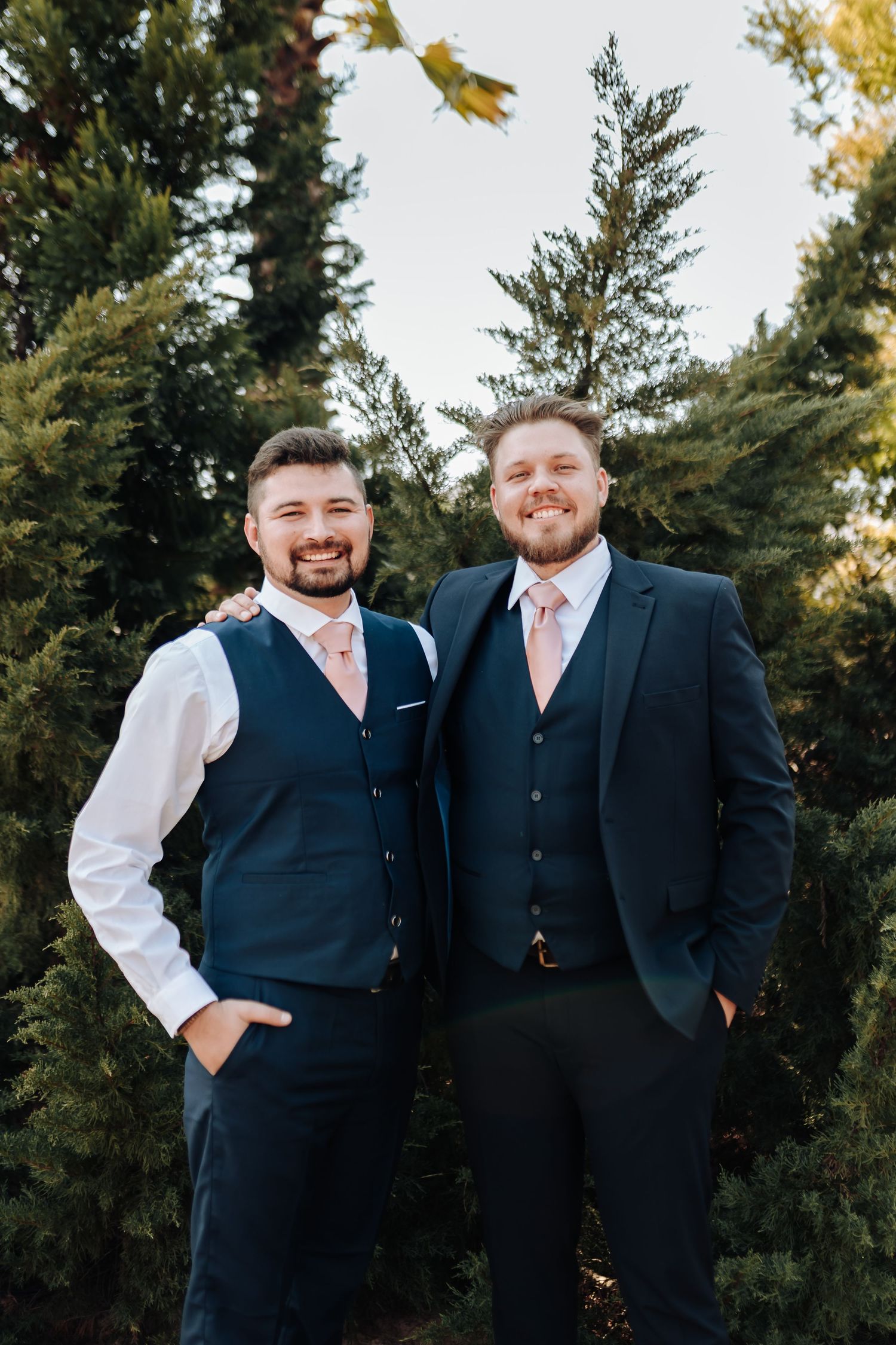 Two people in formal navy suits with pink accents stand together outdoors with evergreen trees in the background.