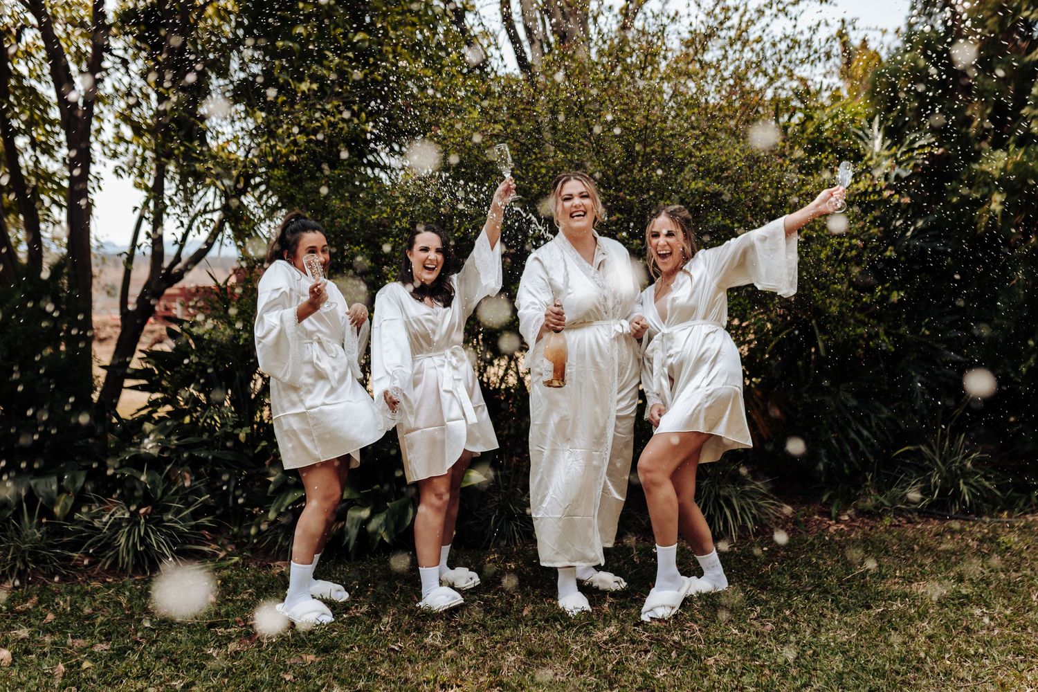 A celebratory group in white robes and fuzzy slippers poses joyfully together in an outdoor garden setting.
