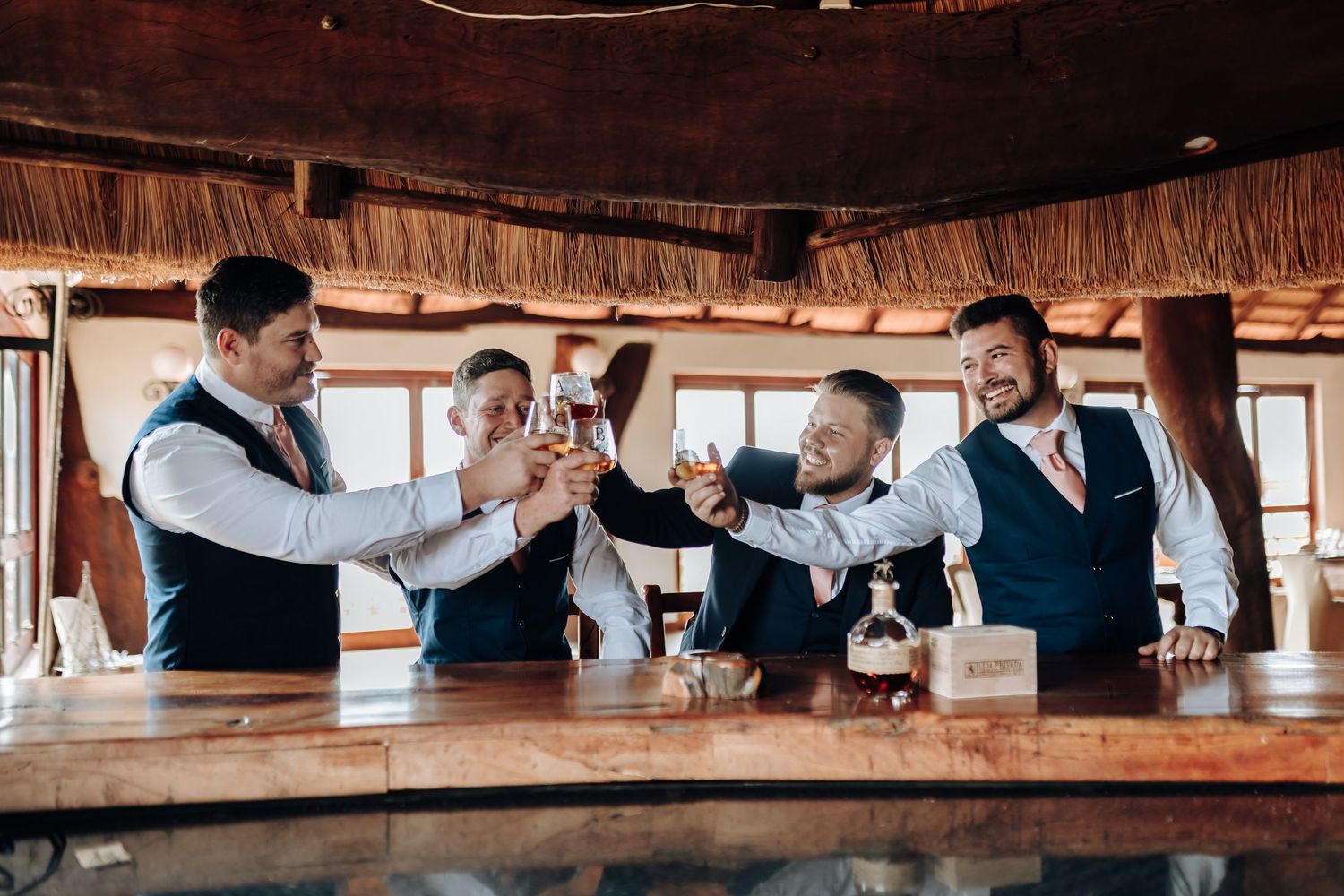 A group of friends in vests raise glasses together at a wooden bar for a celebratory toast.