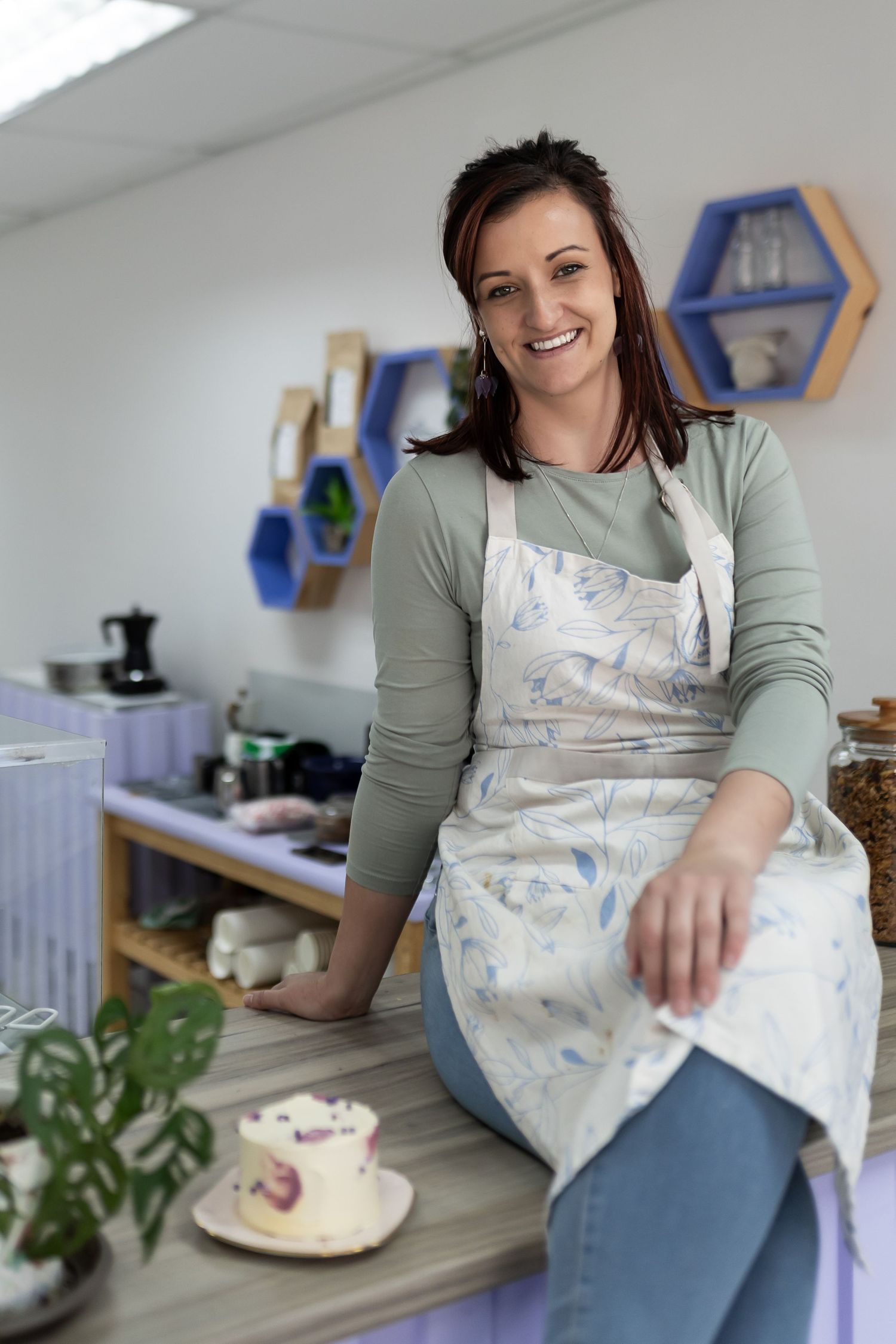 Person in green sweater and patterned apron smiles while preparing food in bright modern kitchen with blue shelving.