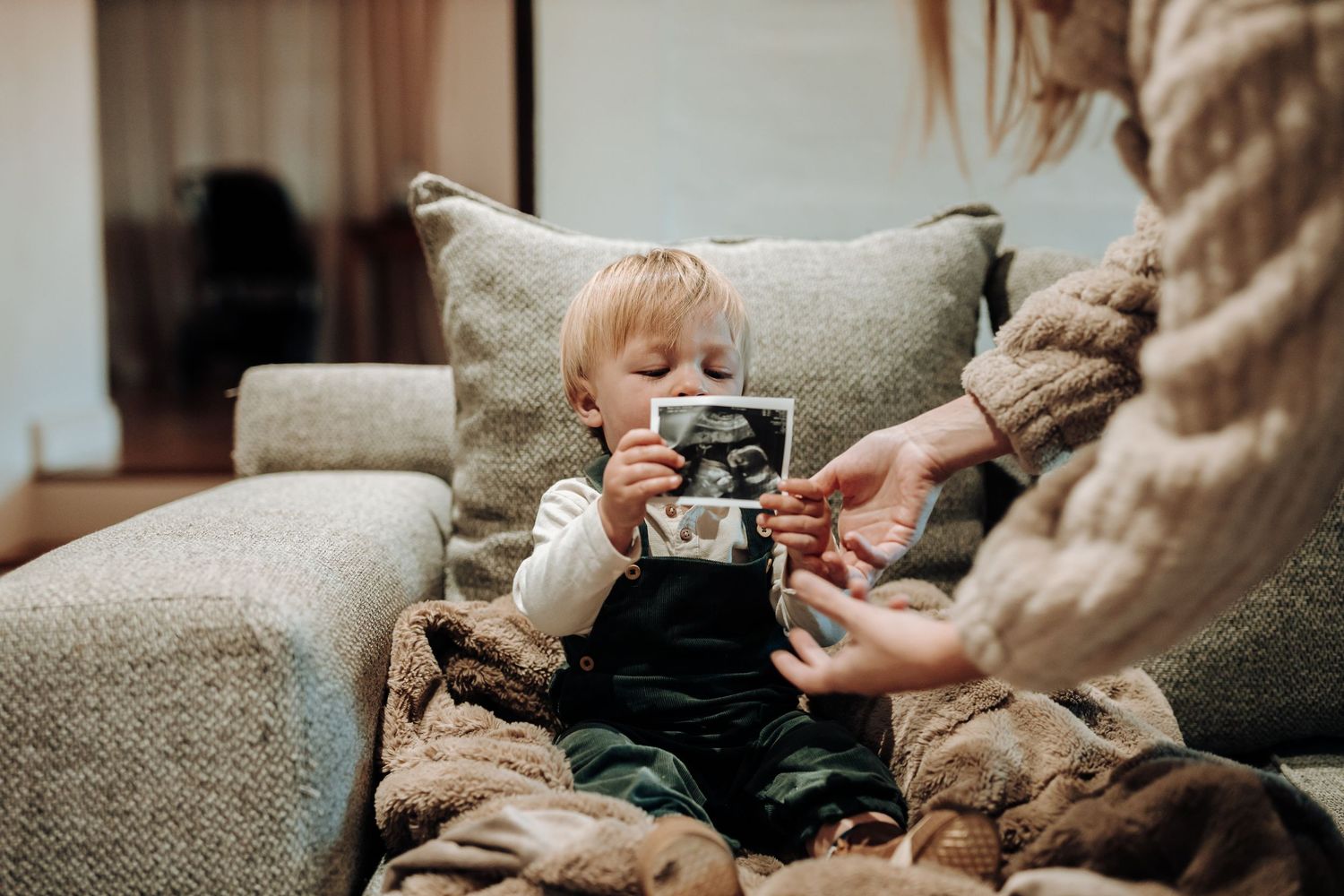 A small child sits on a couch holding a vintage-style camera while someone assists nearby.