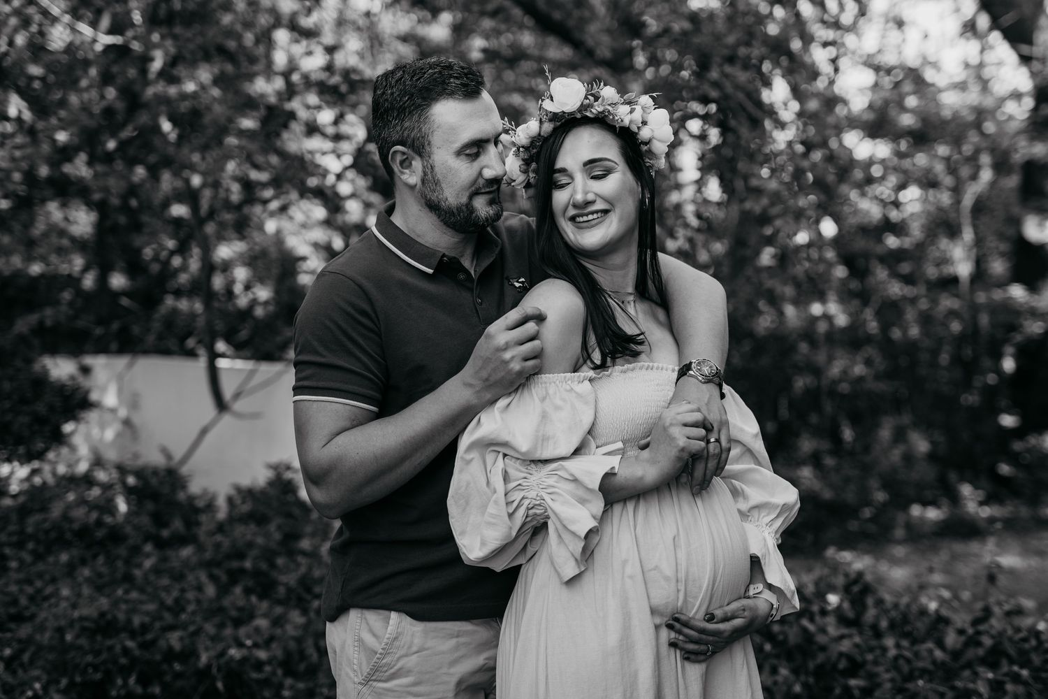 A couple embraces outdoors in black and white as they share a tender moment surrounded by trees.