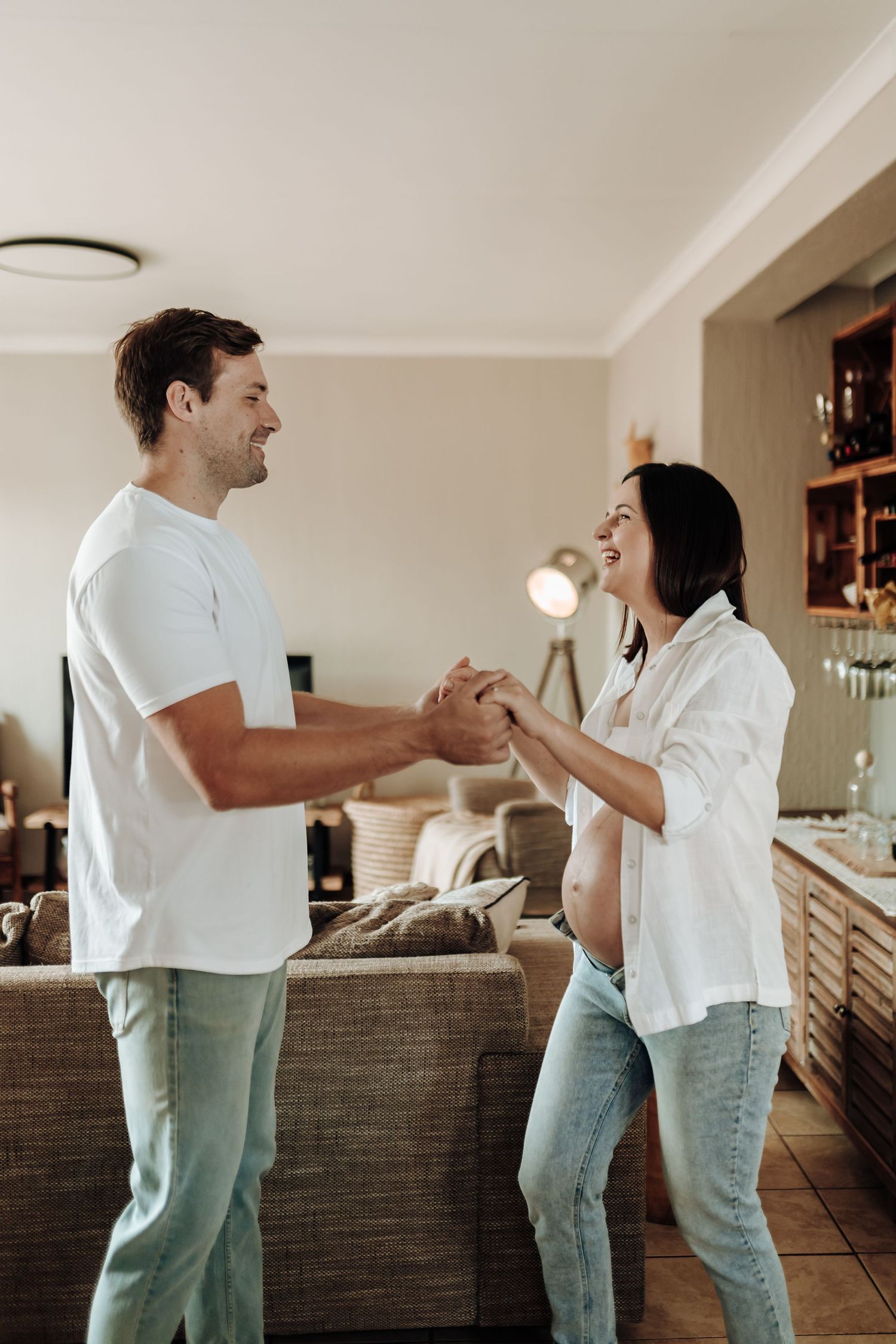 A couple in casual white tops and jeans dances playfully together in their living room.