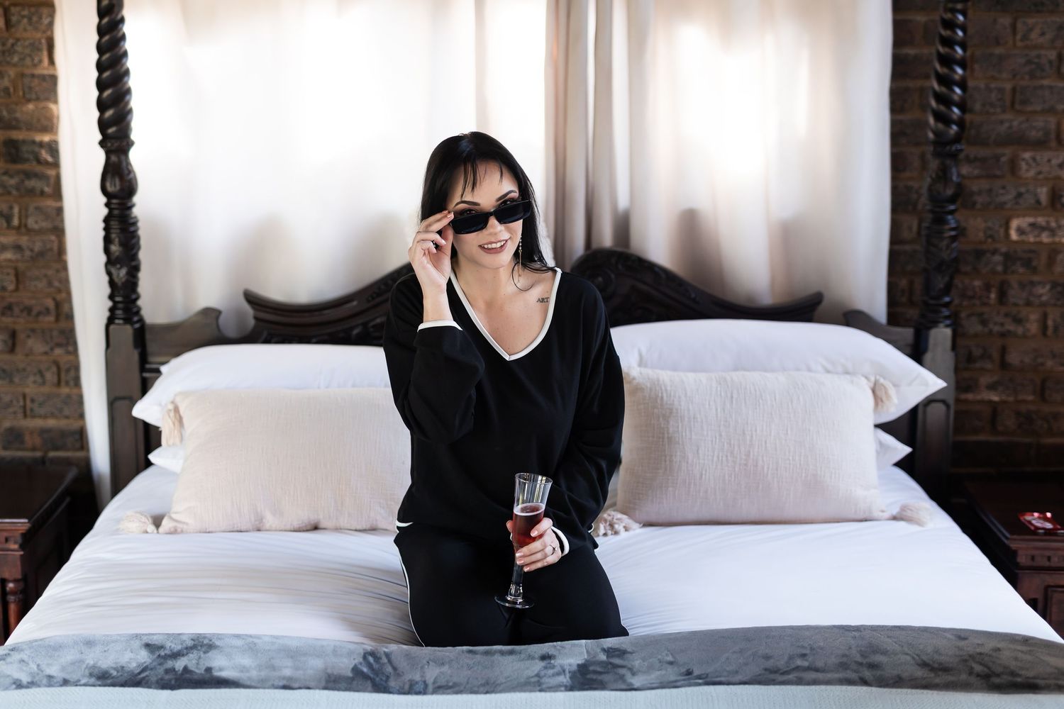 Person in dark clothing and sunglasses sits on bed while holding a beverage in stylish loft bedroom setting.
