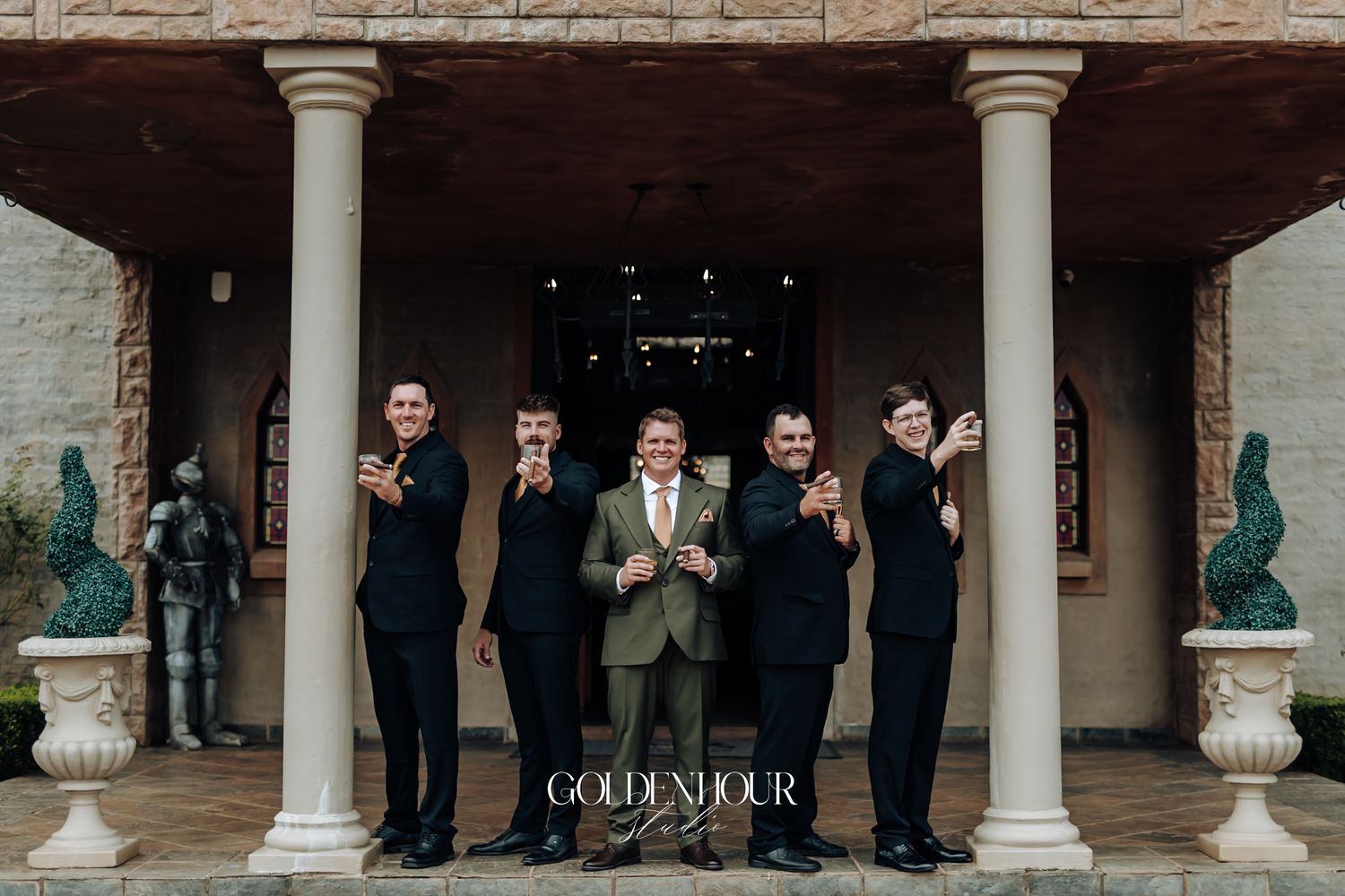 Groom and groomsmen in dark suits posing playfully outside a grand venue with tall columns.
