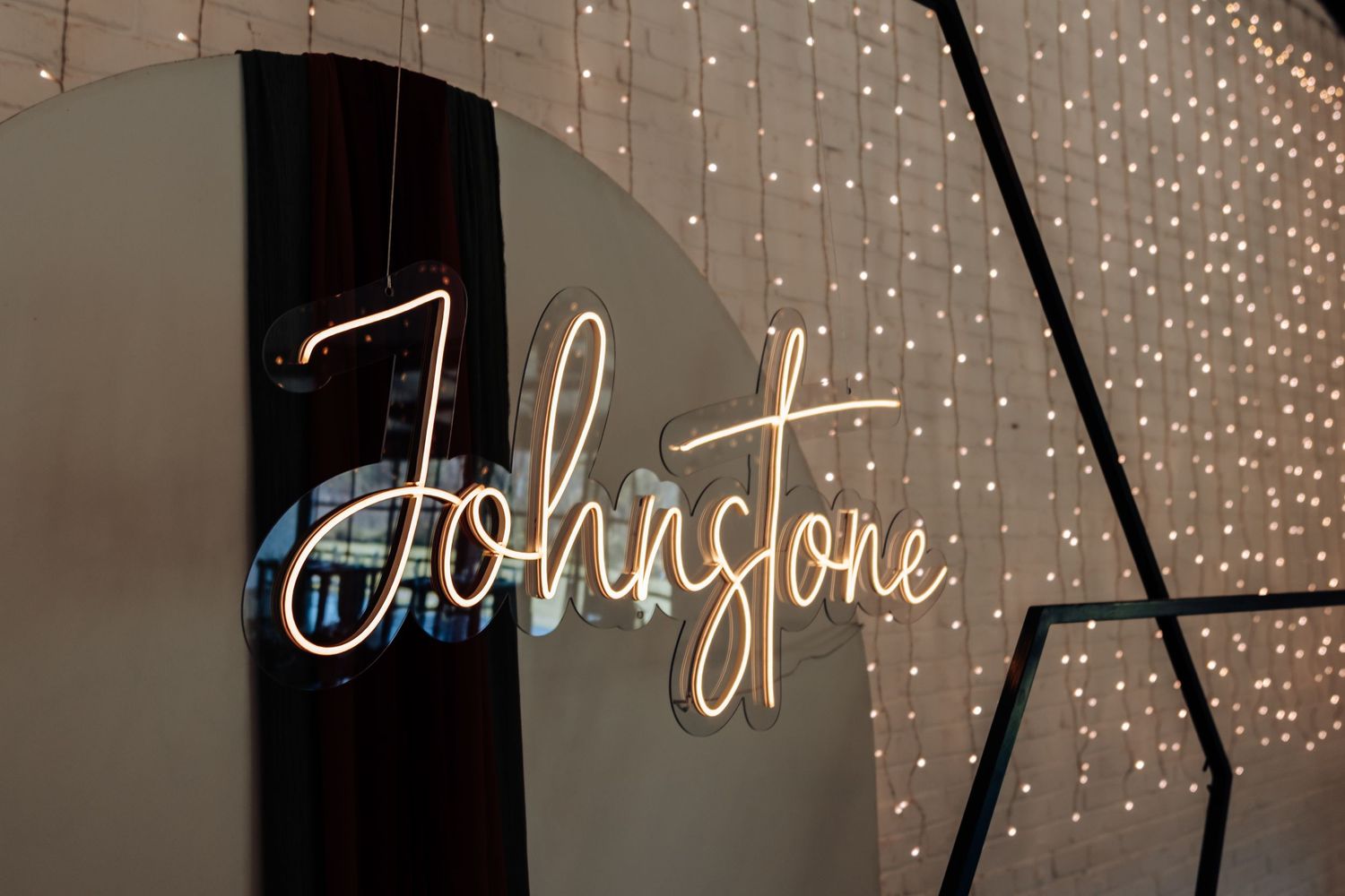 Glowing neon sign spelling Johnstone in cursive script against a backdrop of warm fairy lights.