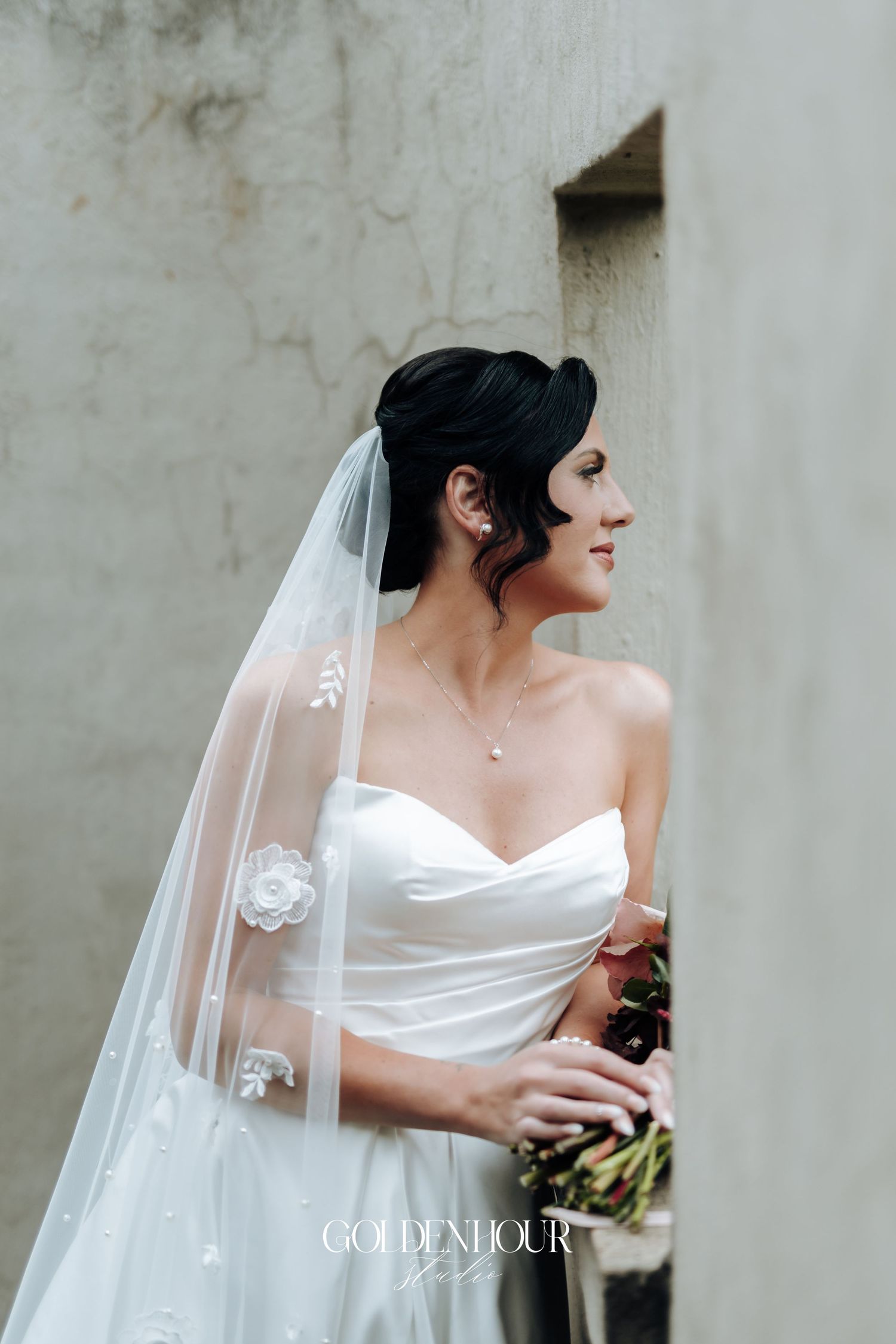 Elegant bride in a strapless white gown and floral veil holding a bouquet beside a textured stone wall.