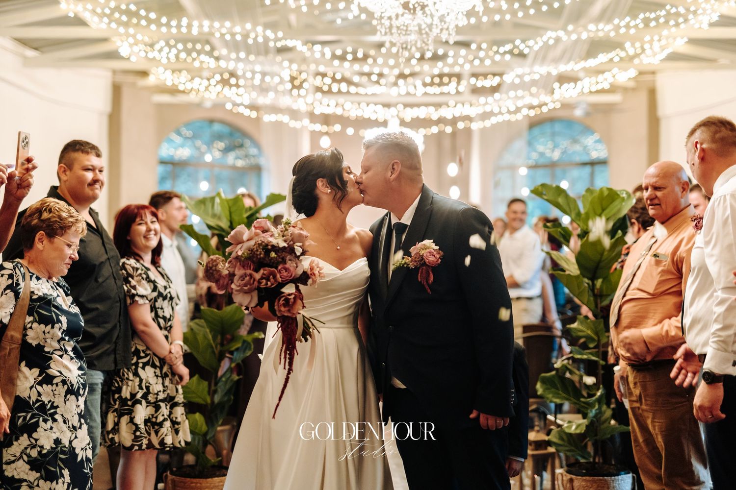Newlyweds share first kiss at altar surrounded by guests under sparkling fairy lights in elegant venue.