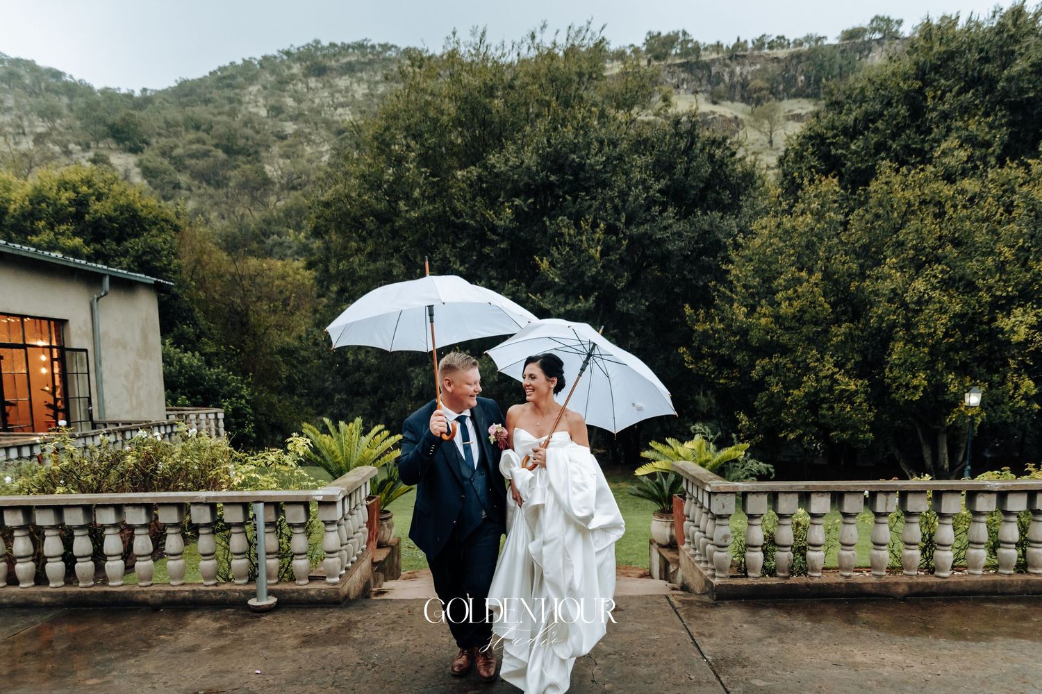 Bride and groom walking with white umbrellas on a rainy day with lush green mountains in the background.