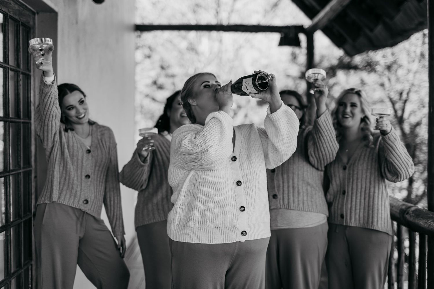 Black and white photo of women celebrating on a porch, one drinking from a bottle while others hold glasses.