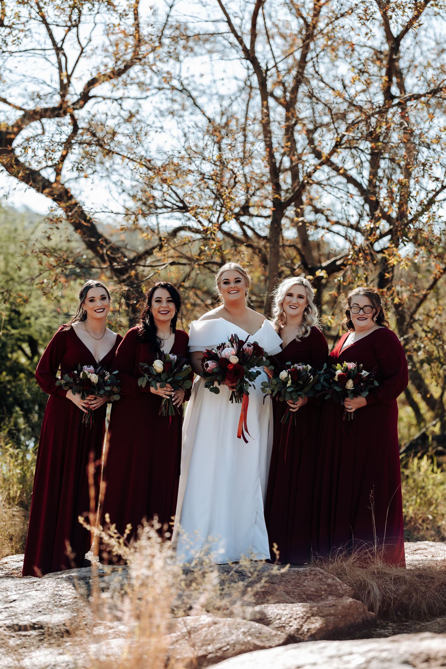 Bride in white gown with four bridesmaids in burgundy velvet dresses holding floral bouquets outdoors.