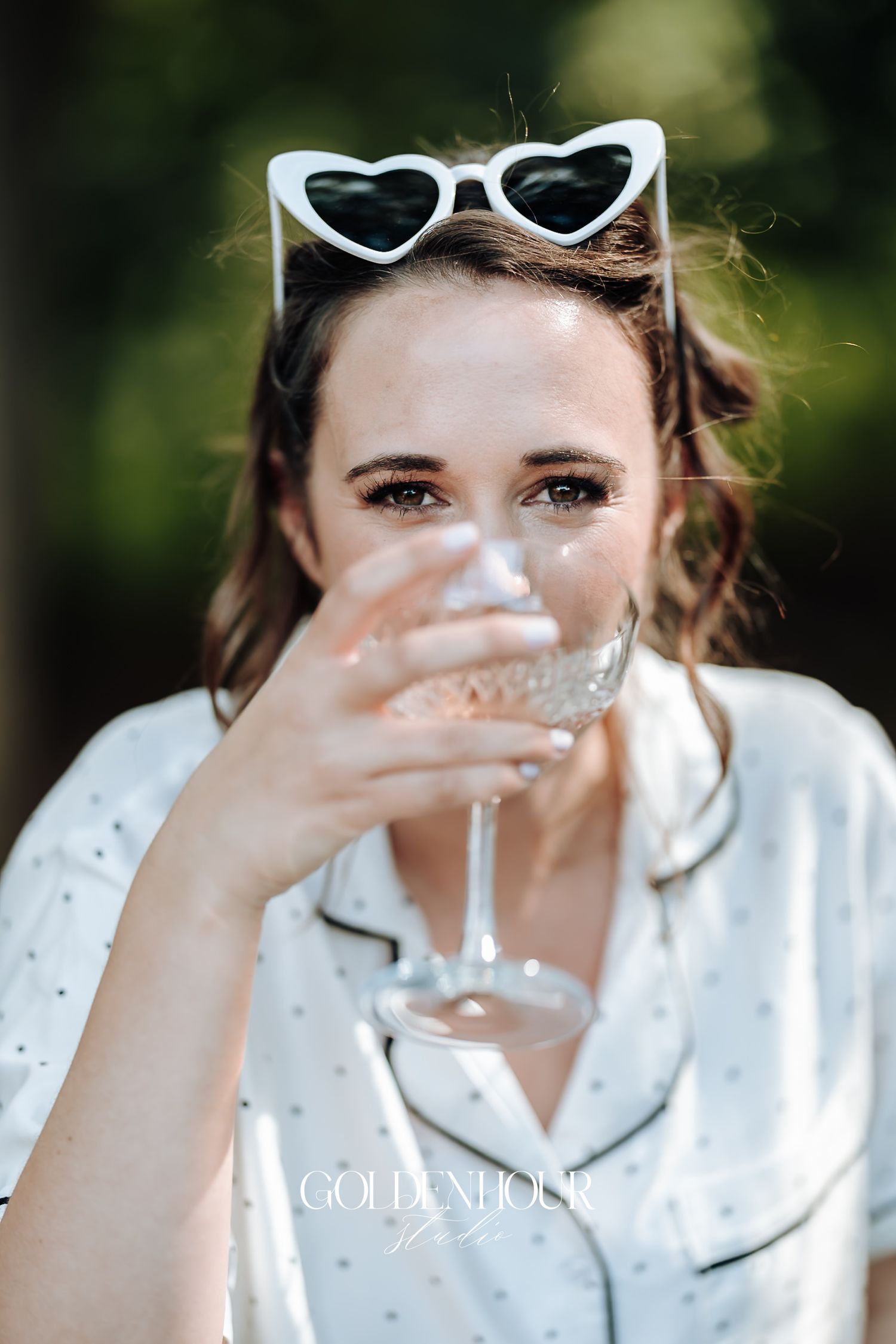 Close-up of bride in polka dot pajamas with white heart sunglasses sipping champagne outdoors.