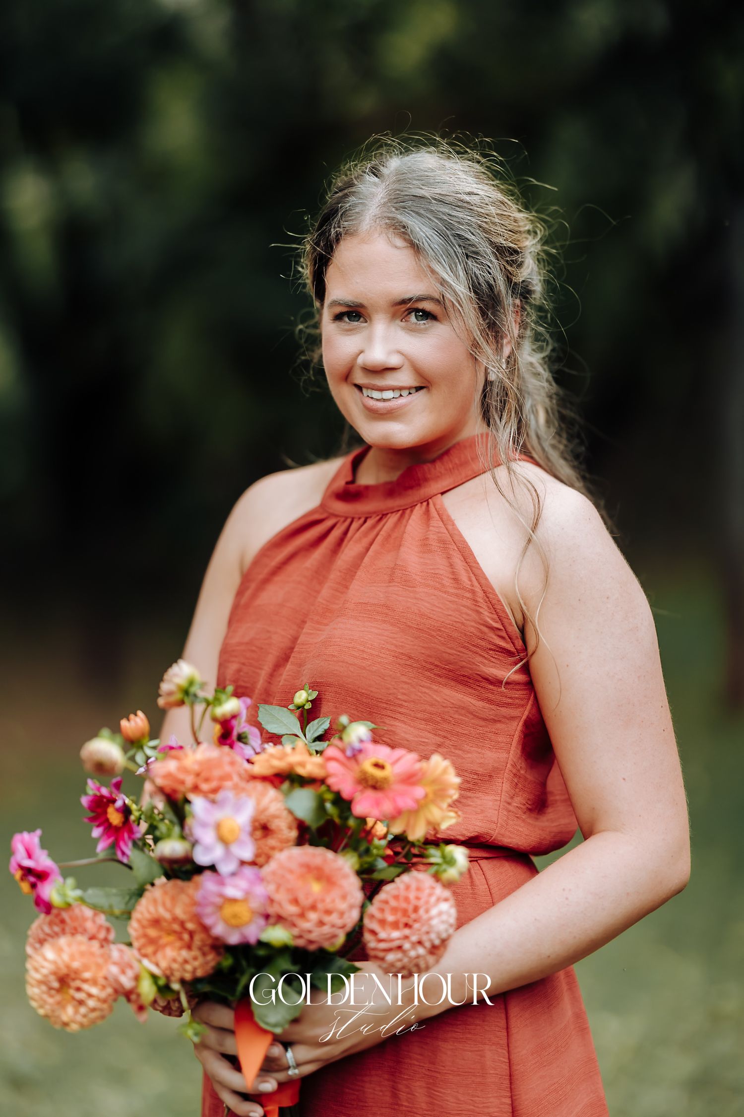 Bridesmaid in rust orange halter dress smiles while holding a peach and pink dahlia bouquet outdoors.
