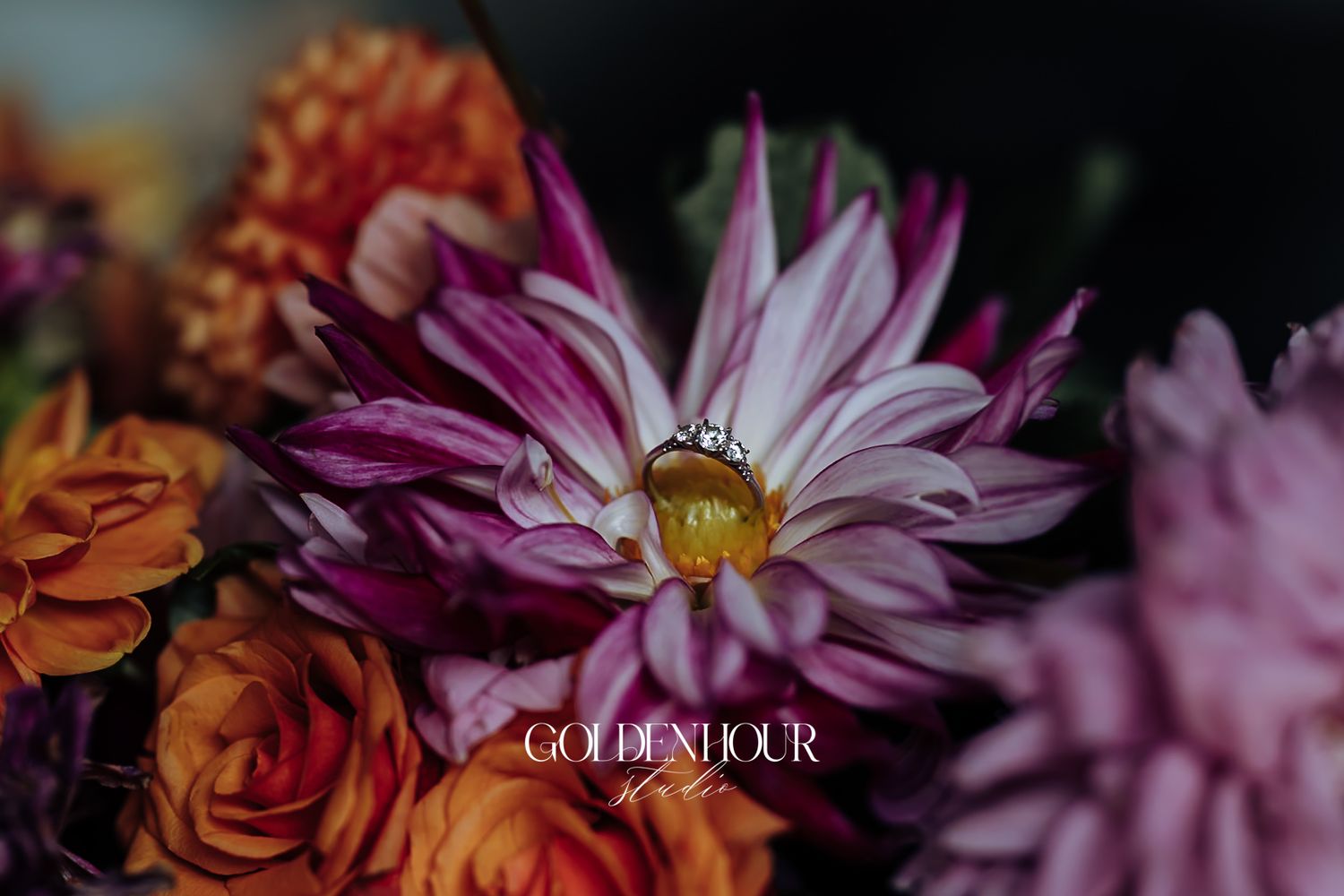 A diamond engagement ring resting on a pink dahlia surrounded by vibrant orange and purple flowers.