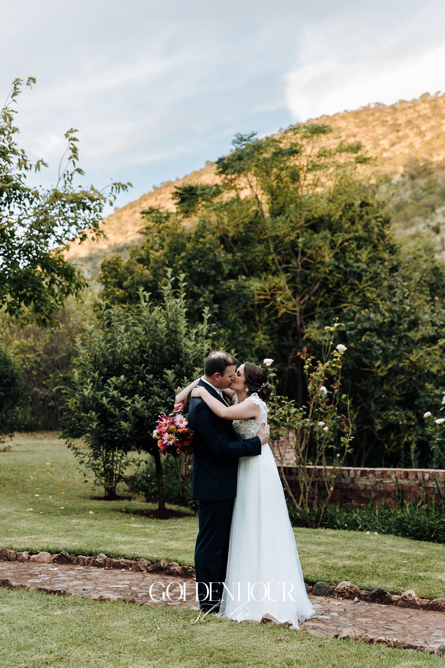 Newlyweds sharing a romantic kiss in a lush garden with mountain backdrop.