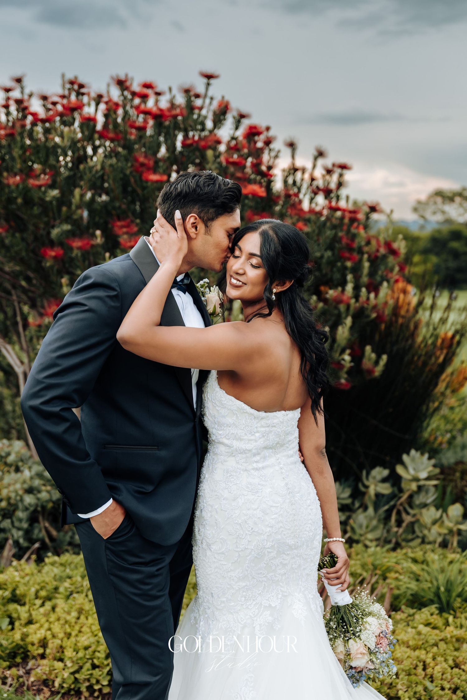 Romantic wedding portrait in garden with red flowering bush and cloudy sky.