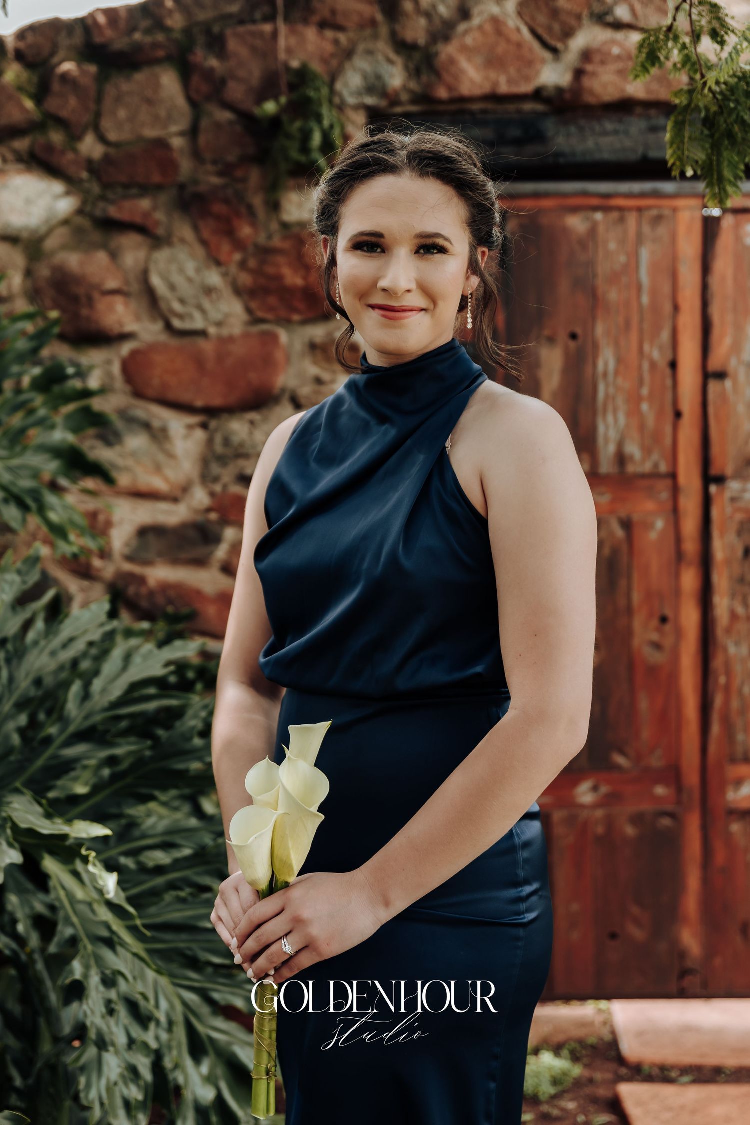 Elegant navy blue one-shoulder dress photographed against rustic brick wall and greenery backdrop.