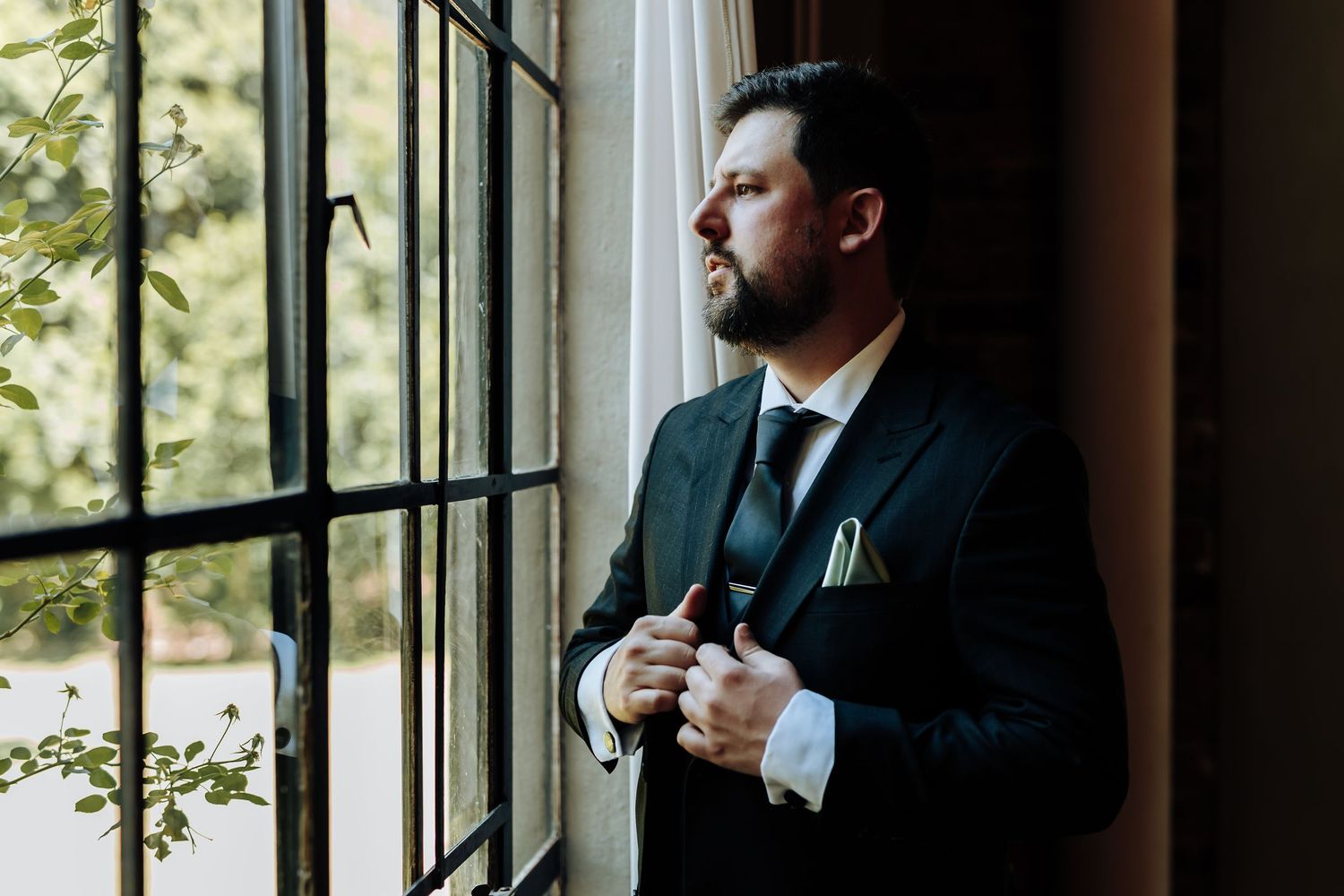 Men in dark suits getting ready by adjusting their ties near large windows with natural light.