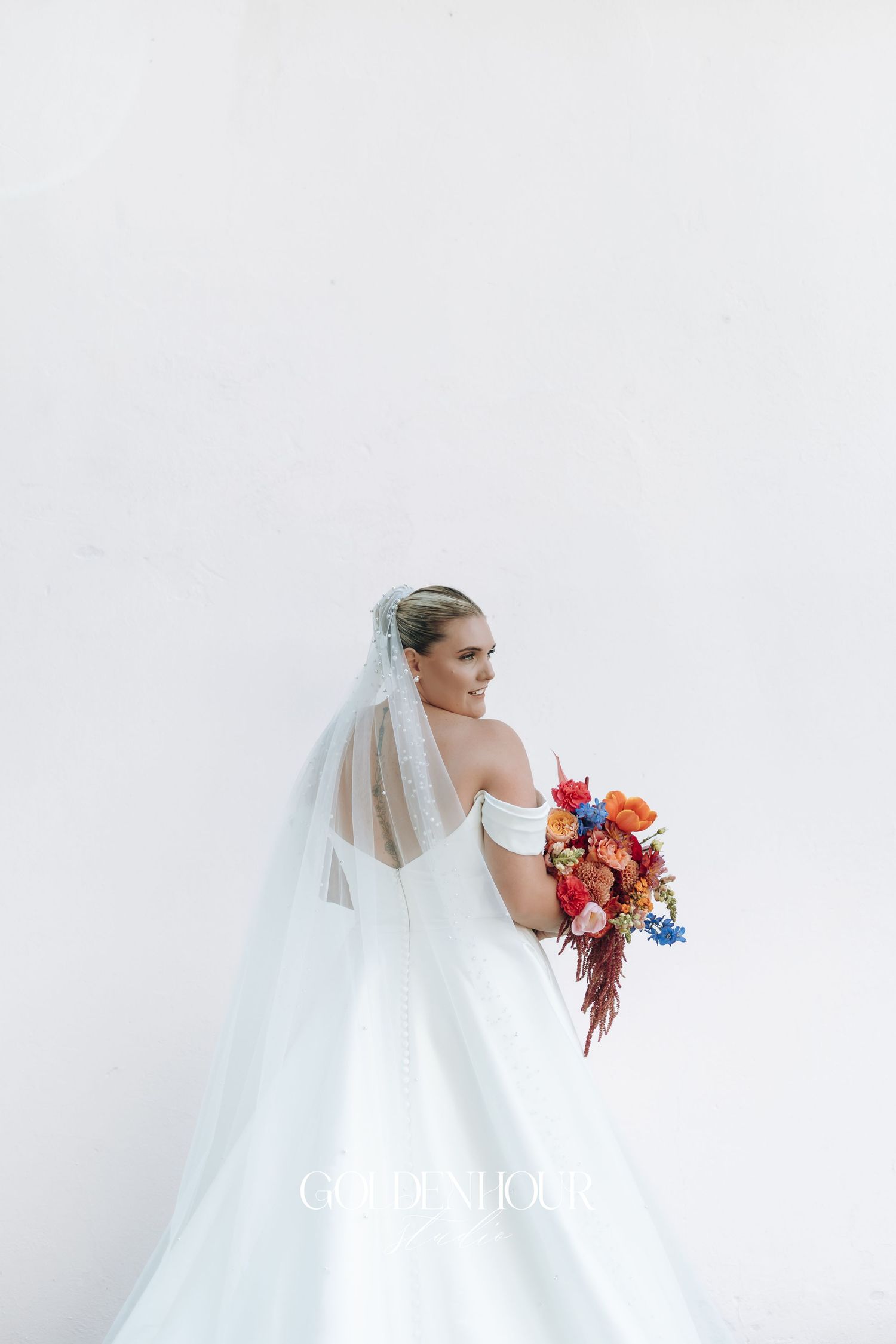 A bride in a flowing white wedding gown holds a vibrant bouquet of red and blue flowers against a light background.