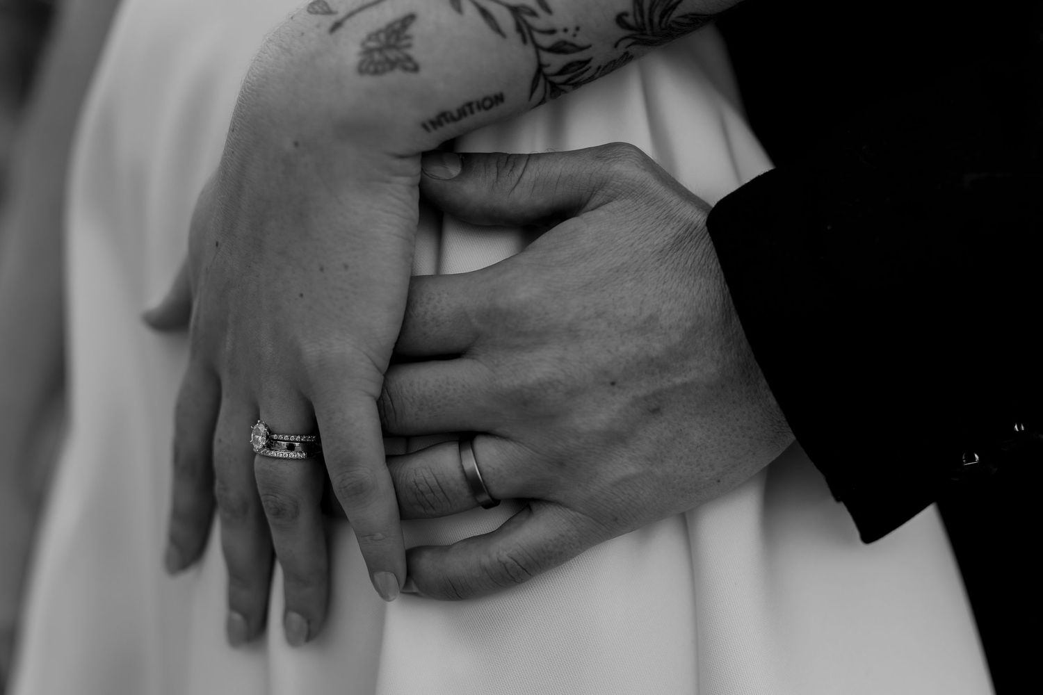 Black and white closeup of hands with wedding rings embracing around white dress fabric.
