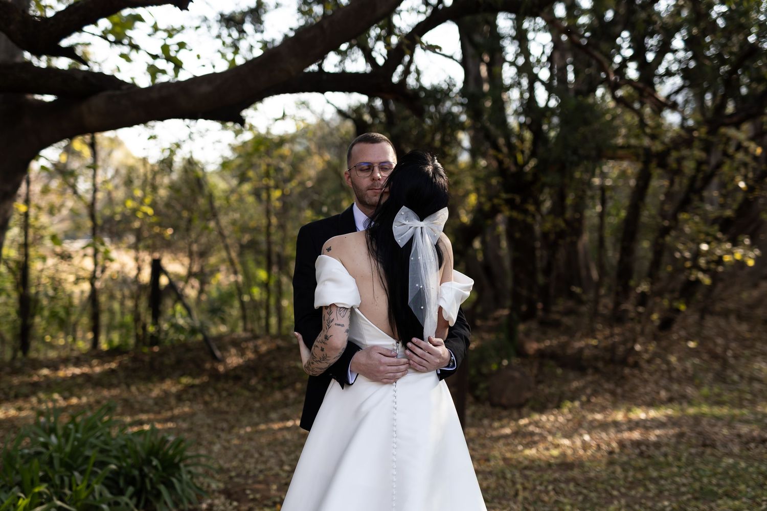 A couple in wedding attire embraces in a wooded outdoor setting during golden hour.