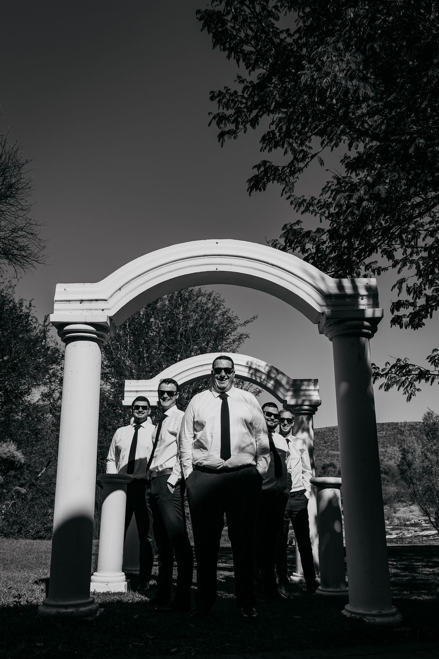 Black and white photo of groomsmen in white shirts and ties posing under a stone arch outdoors.