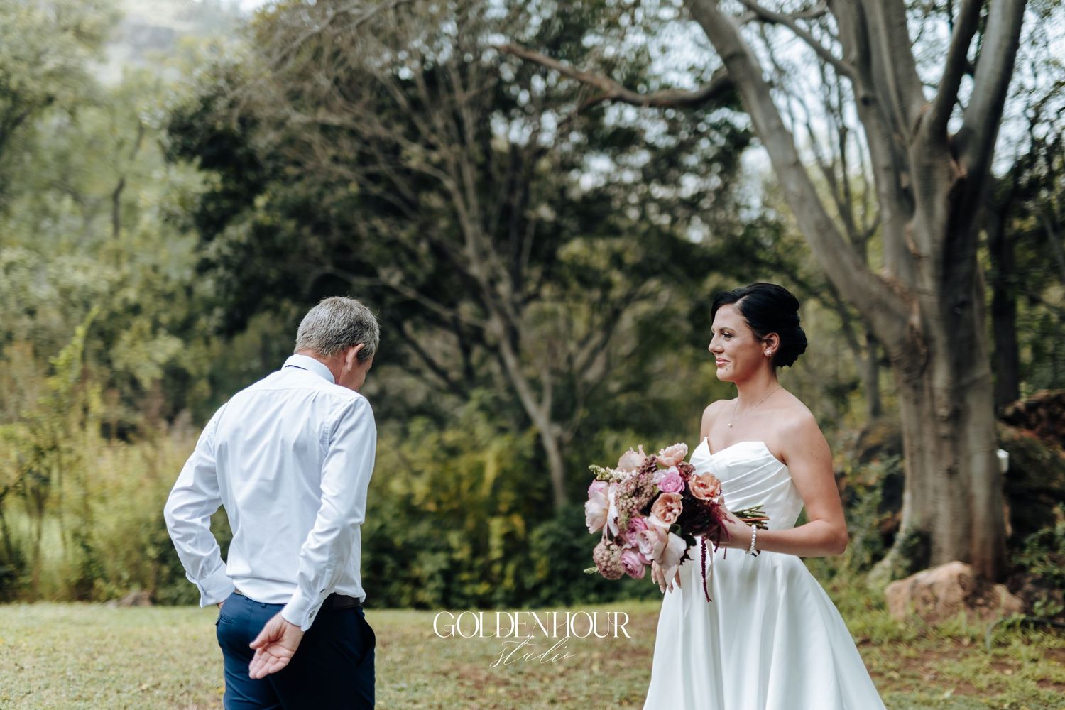 Bride in white strapless gown holding pink bouquet during outdoor first look moment with groom in lush garden setting.