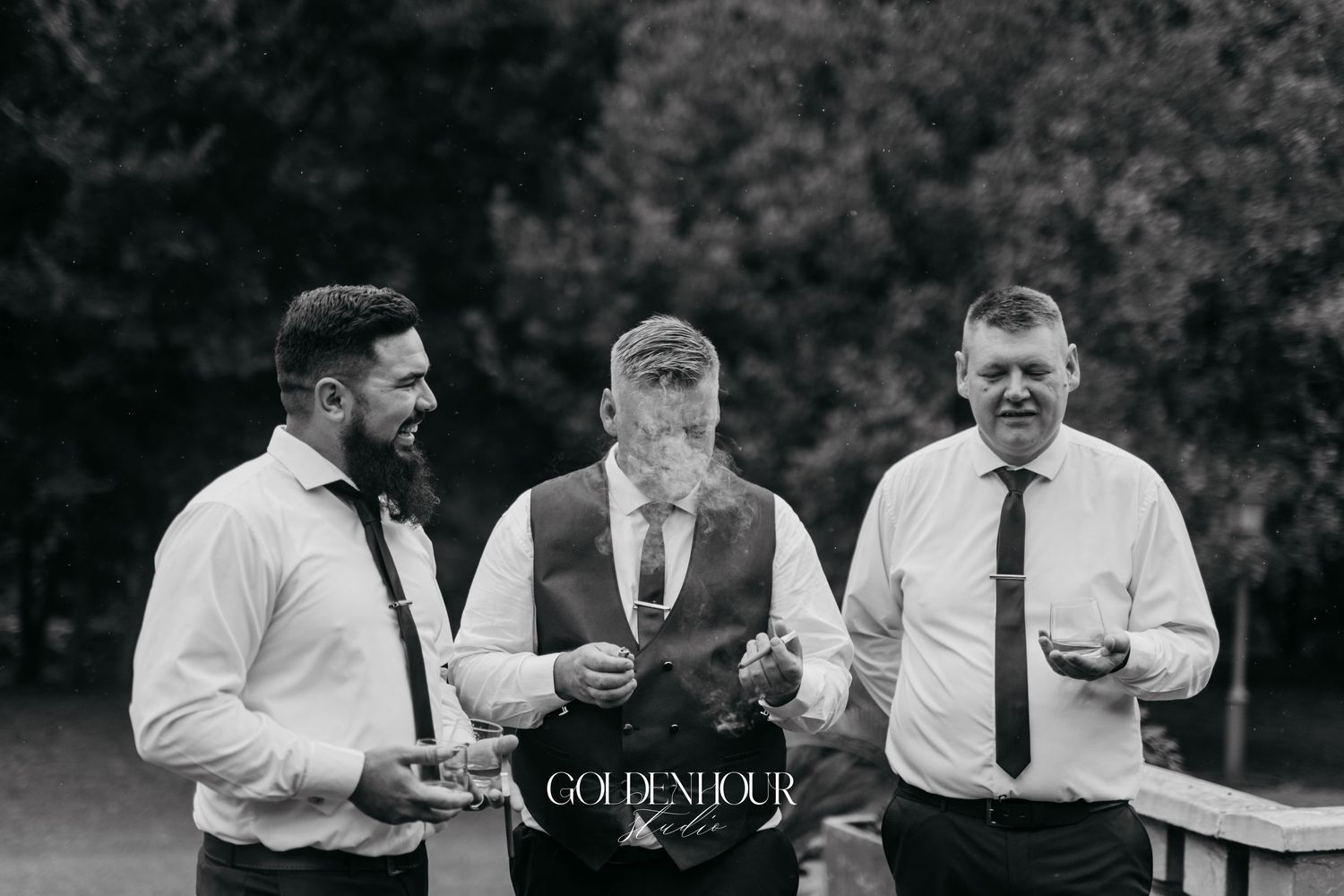 Black and white photo of three groomsmen laughing and chatting outdoors at a wedding celebration.
