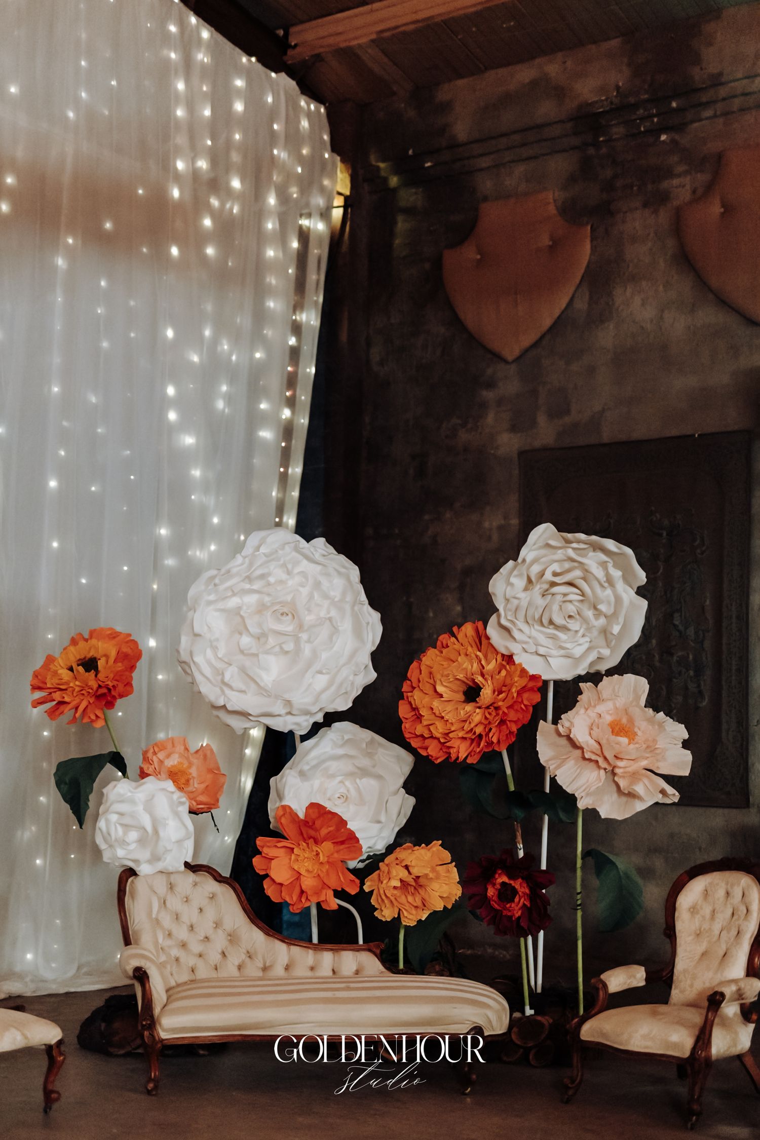 Elegant wedding backdrop with oversized white and orange paper flowers beside a tufted cream chaise lounge with fairy lights.