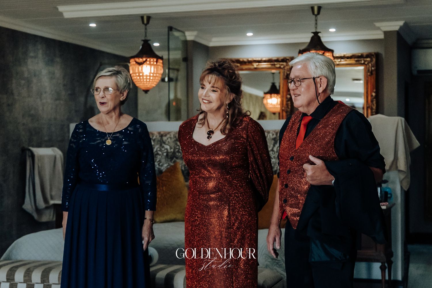 Three elegantly dressed wedding guests pose indoors, two women in blue and red gowns with an older gentleman.