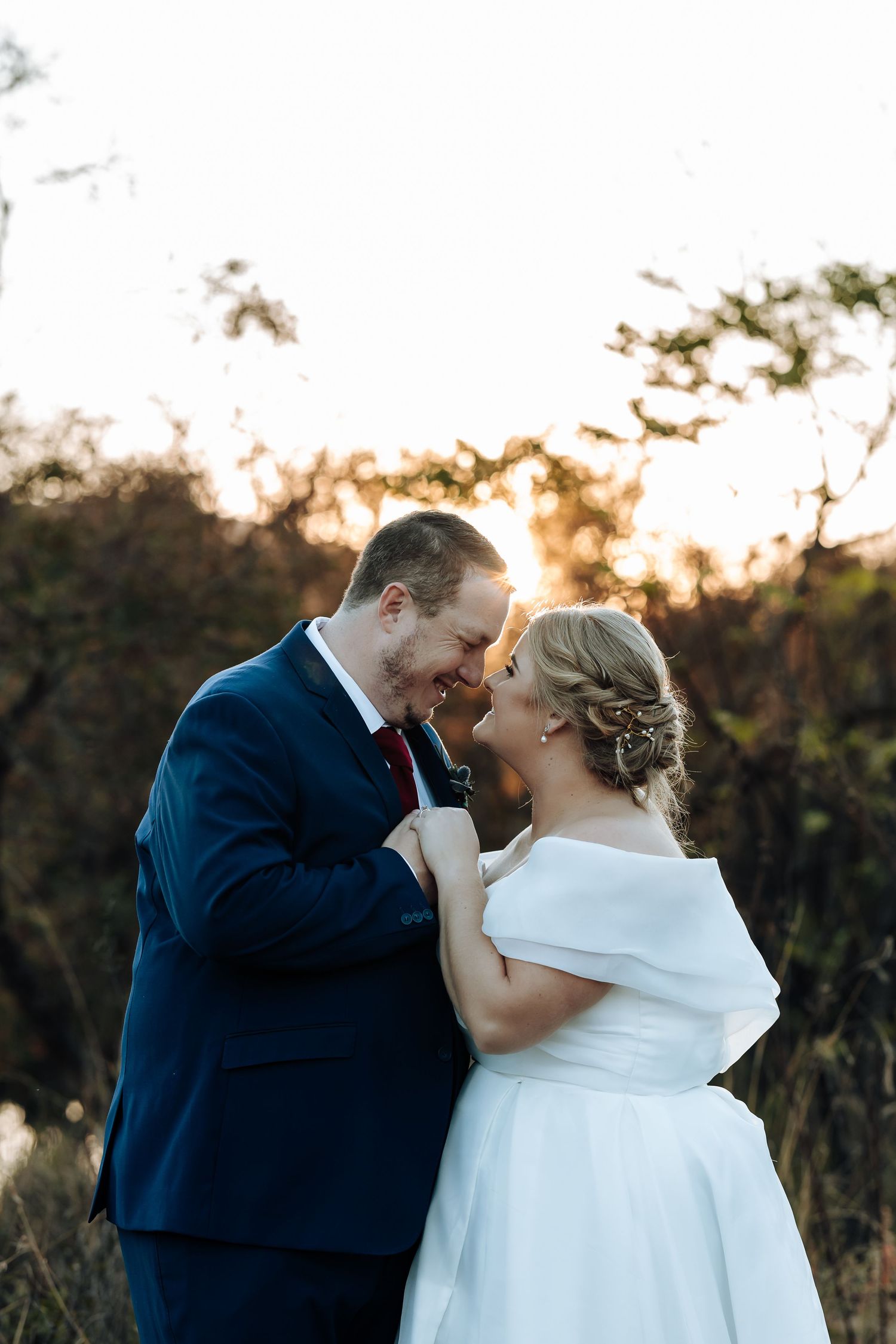 A bride and groom share a romantic forehead-to-forehead moment outdoors at golden hour, she in white off-shoulder gown, he in navy suit.