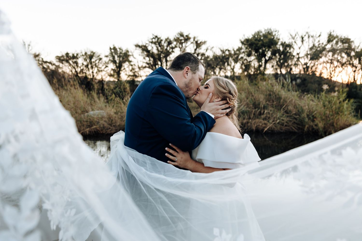Bride and groom share a romantic kiss at sunset with flowing white veil in foreground at outdoor wedding.
