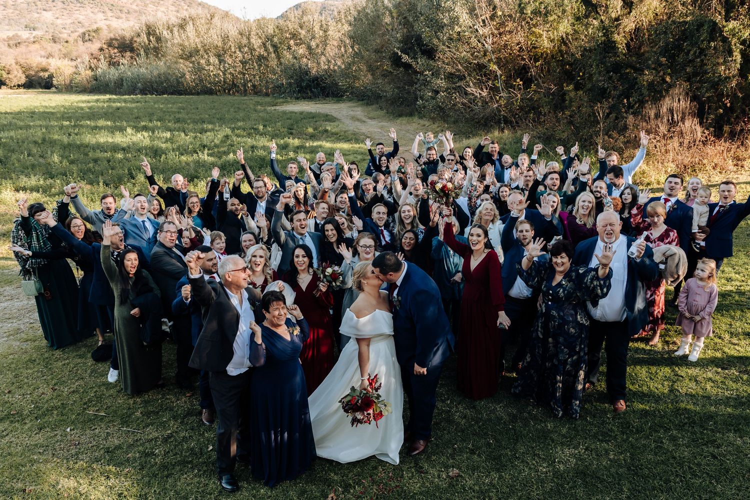 Bride and groom kiss surrounded by large wedding party and guests outdoors in scenic mountain meadow.