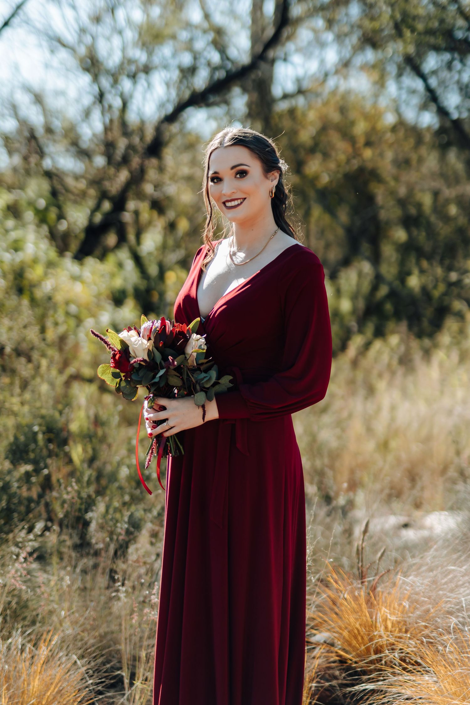 Woman in burgundy velvet gown holding floral bouquet outdoors in natural bush setting.