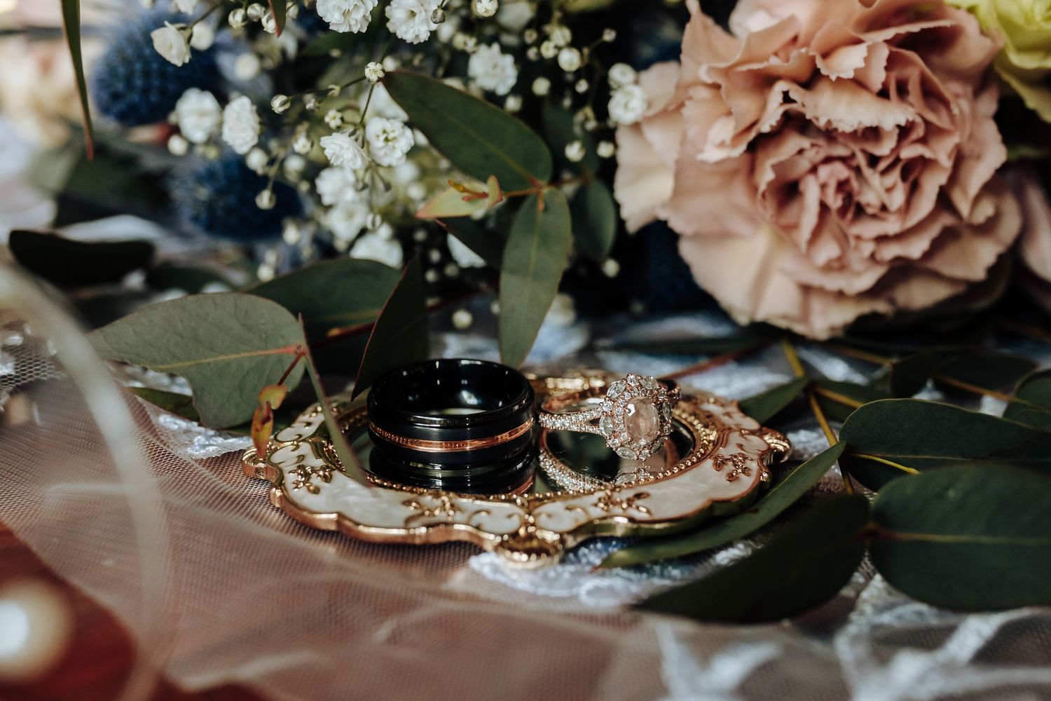 Wedding rings with delicate rose gold jewelry displayed on greenery with blush pink roses and baby's breath flowers.