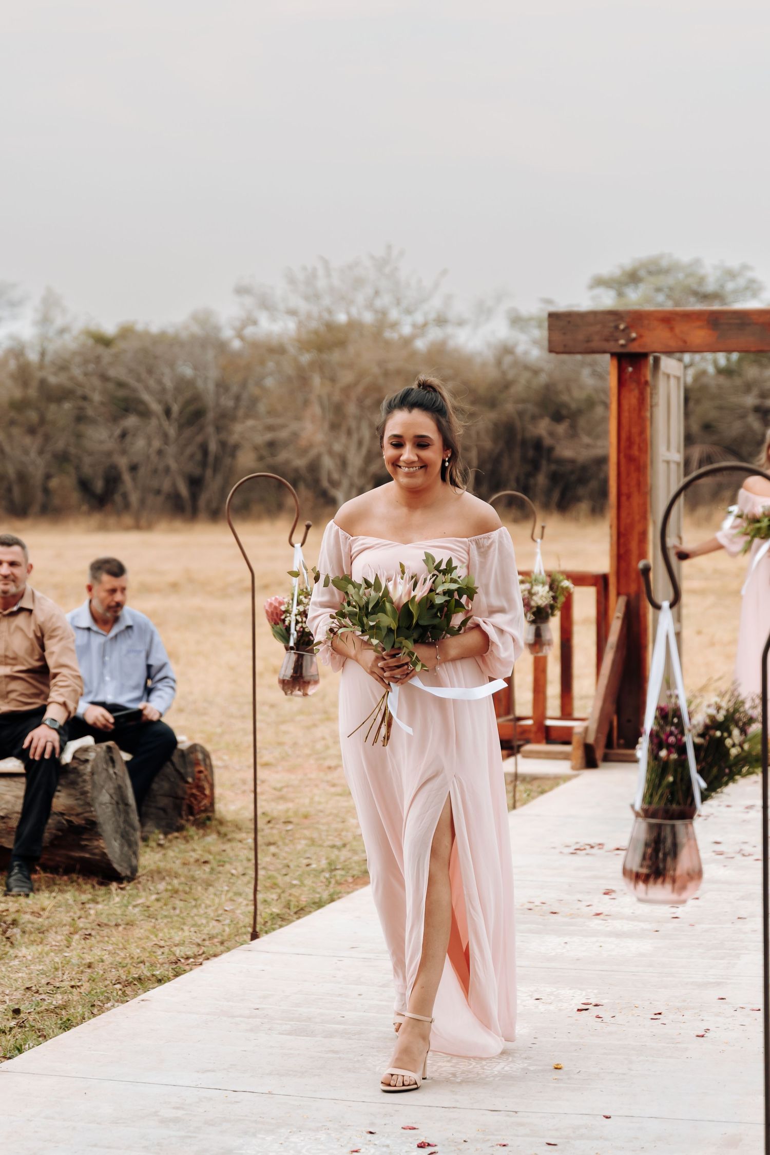 A wedding party member walks down an outdoor aisle in a flowing off-shoulder dress carrying a bouquet of flowers.