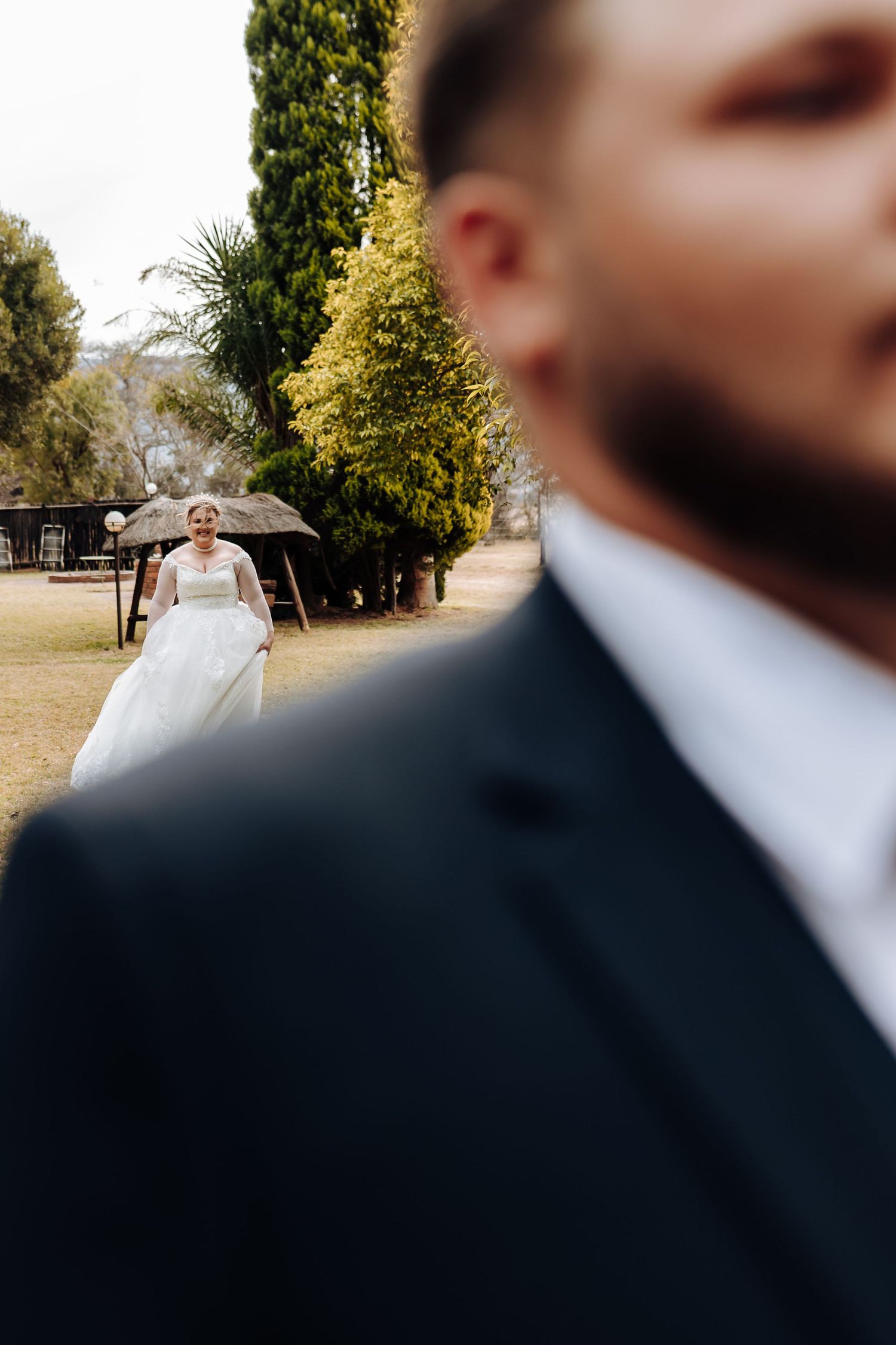 A wedding photograph with soft focus on foreground figure while bride stands in sunny garden background.