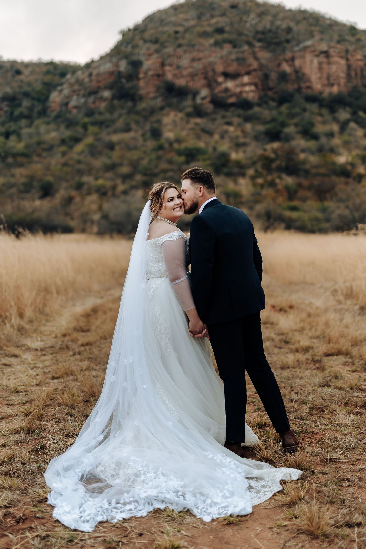 Wedding couple stands in golden field with dramatic mountain backdrop and flowing white lace train.