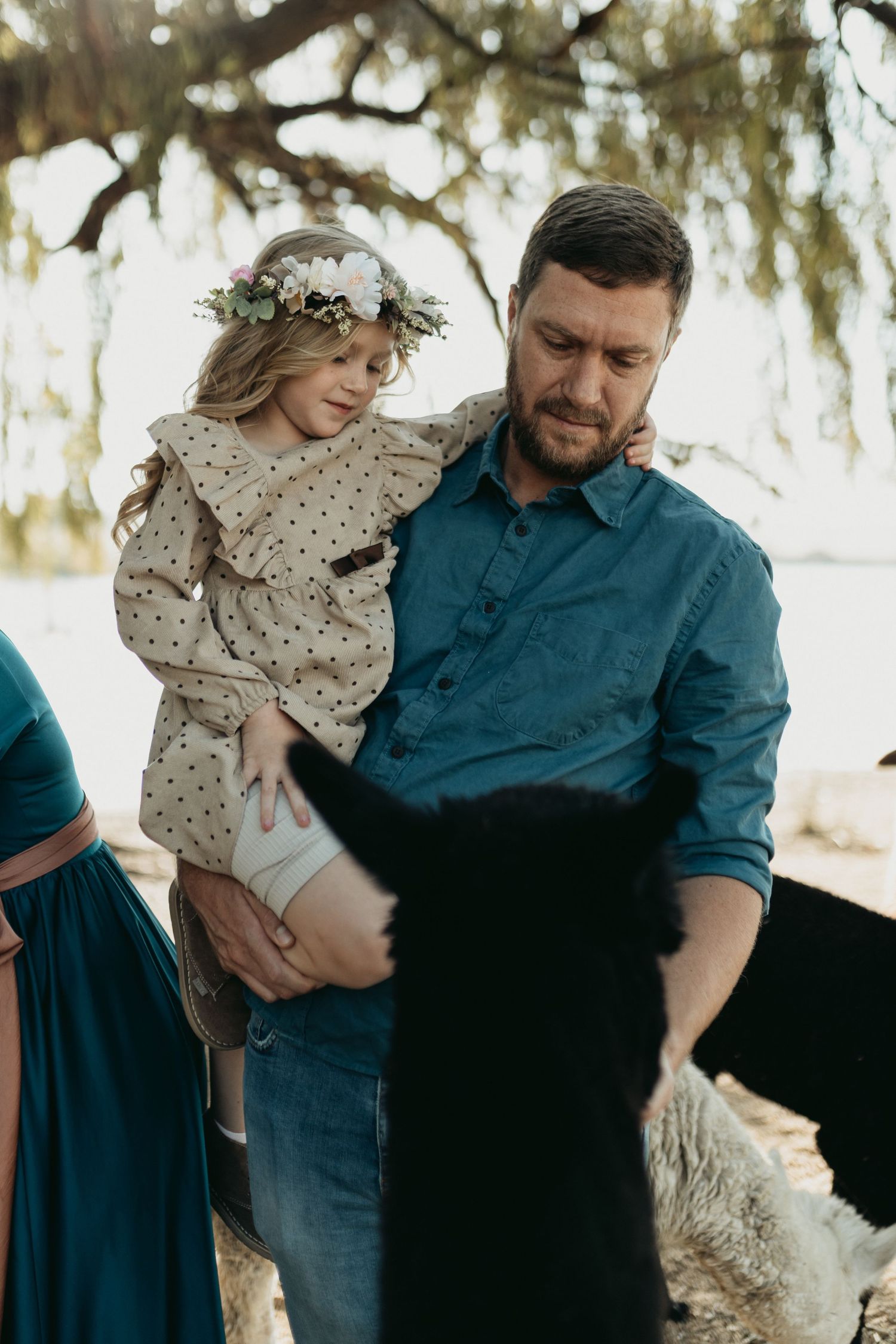 A tender family moment captured outdoors under moss-draped trees with floral crowns and casual attire.