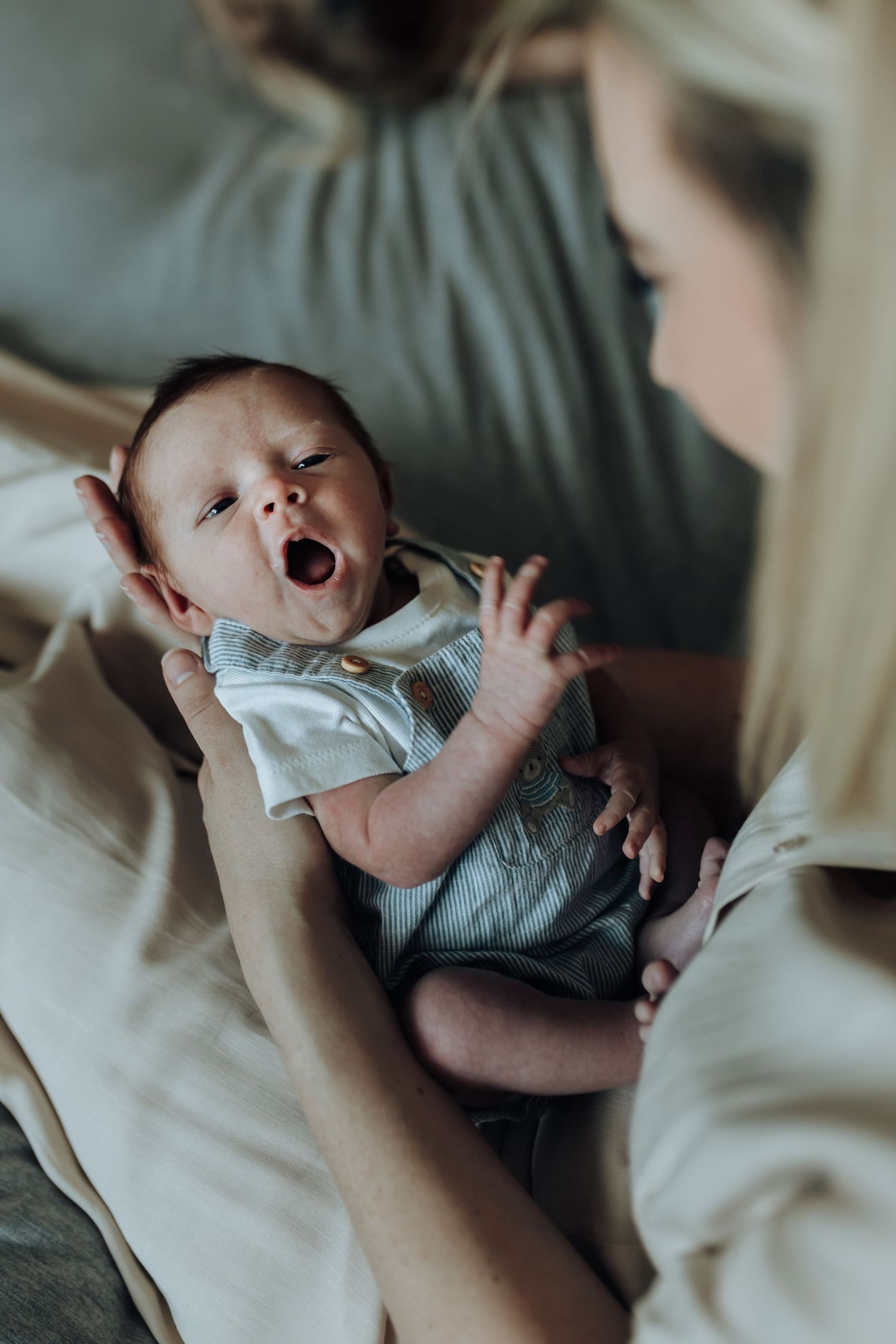 A tiny baby in gray clothing yawns while being held in a soft-focus indoor setting.