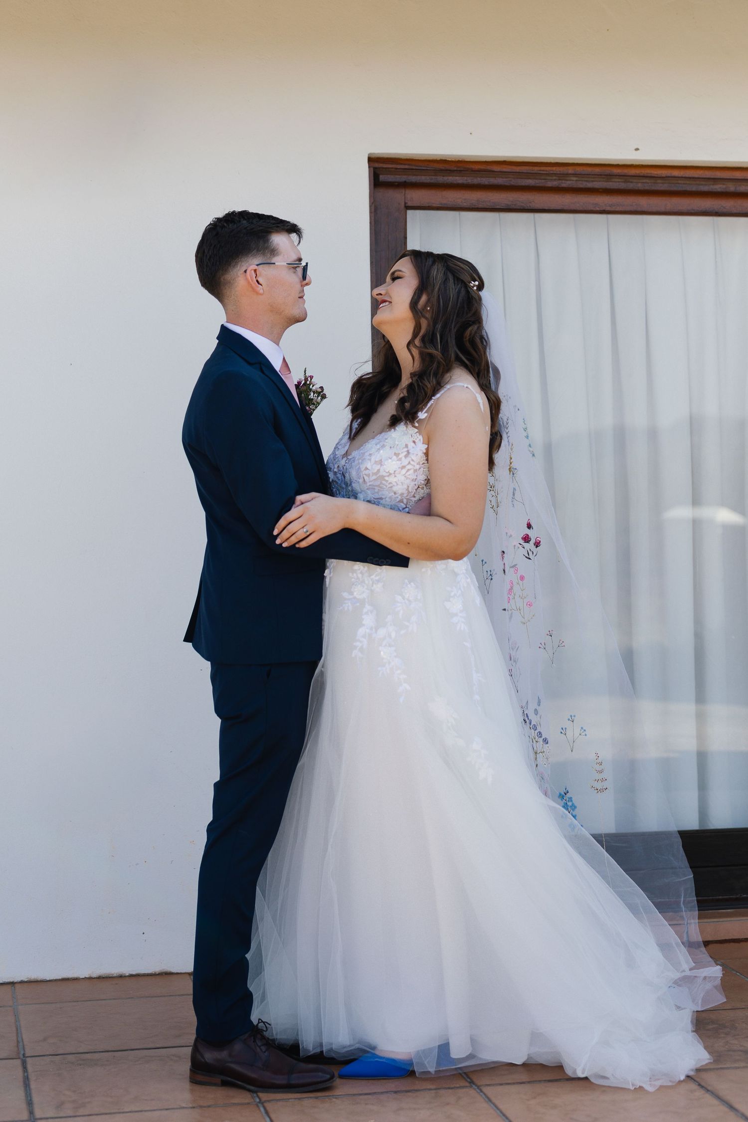 A couple in wedding attire share a tender moment in front of white curtains during their first dance.
