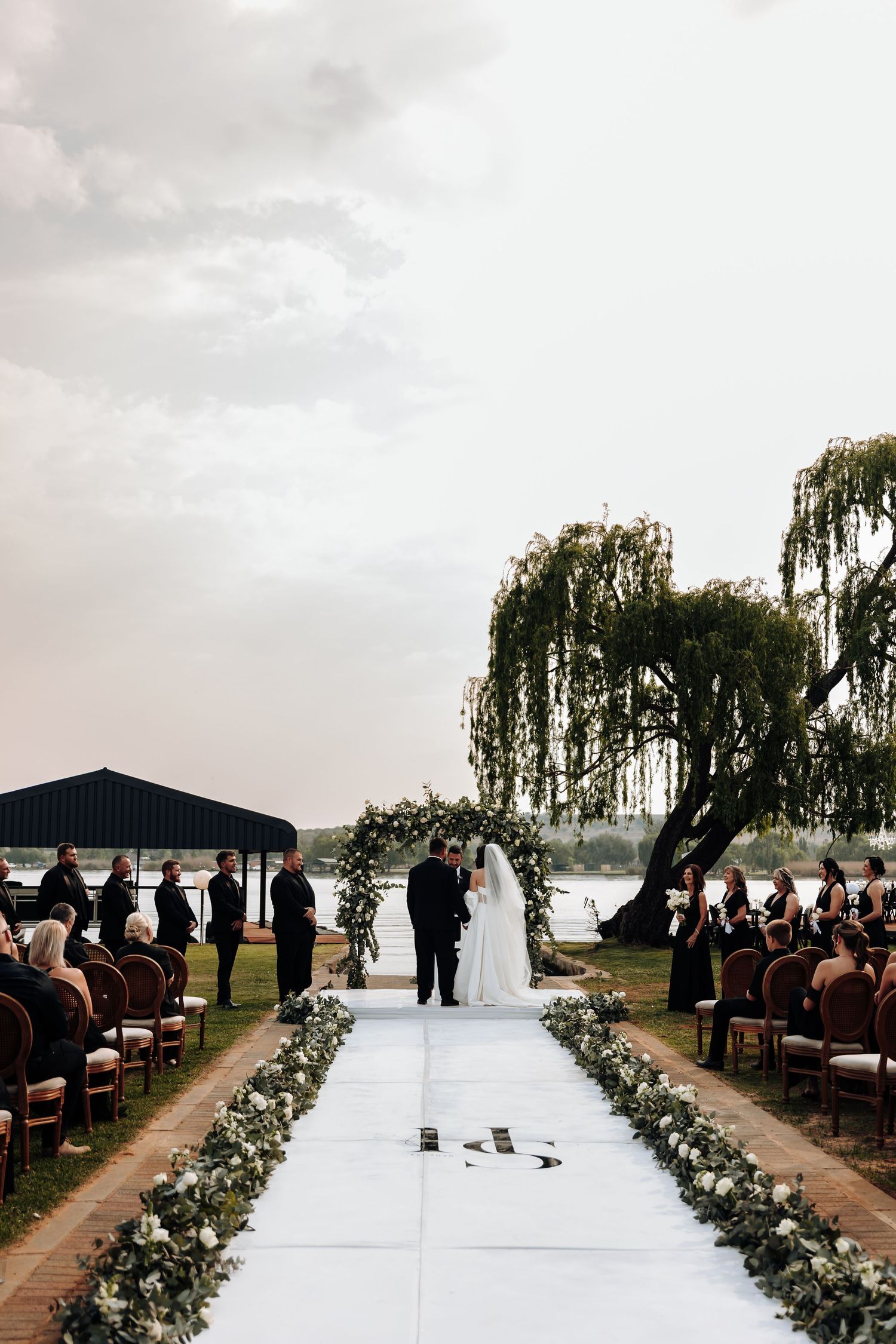 Outdoor wedding ceremony setup with white aisle runner and floral arrangements along the path under weeping willow trees.