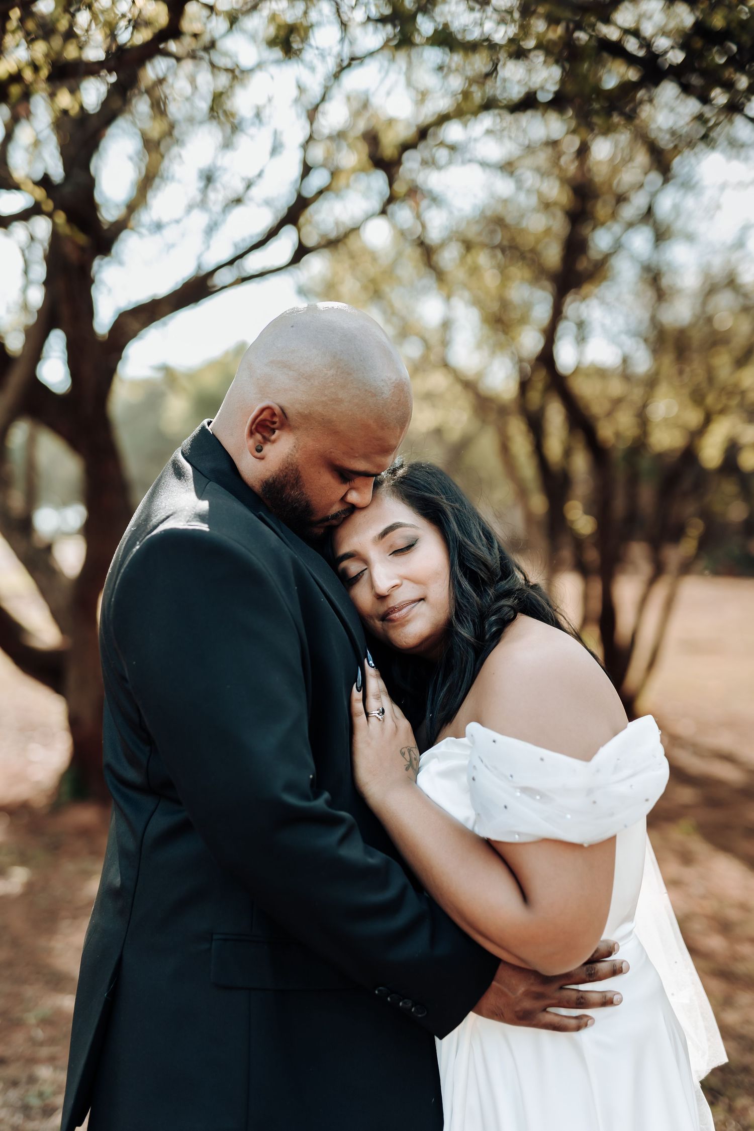 Tender moment between newlyweds embracing under trees in natural outdoor setting.