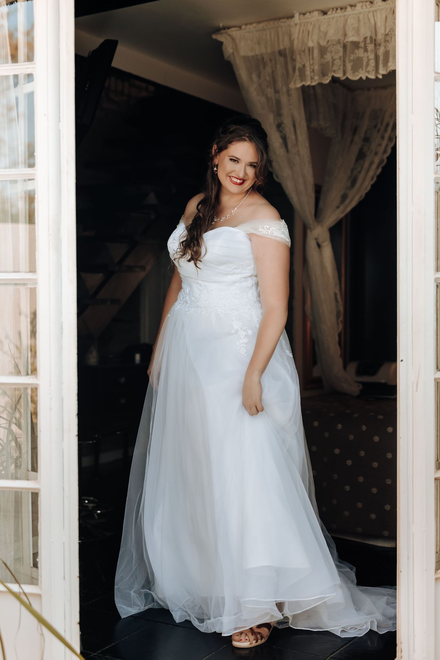 A bride in an elegant off-shoulder wedding dress stands in a doorway with lace curtains.