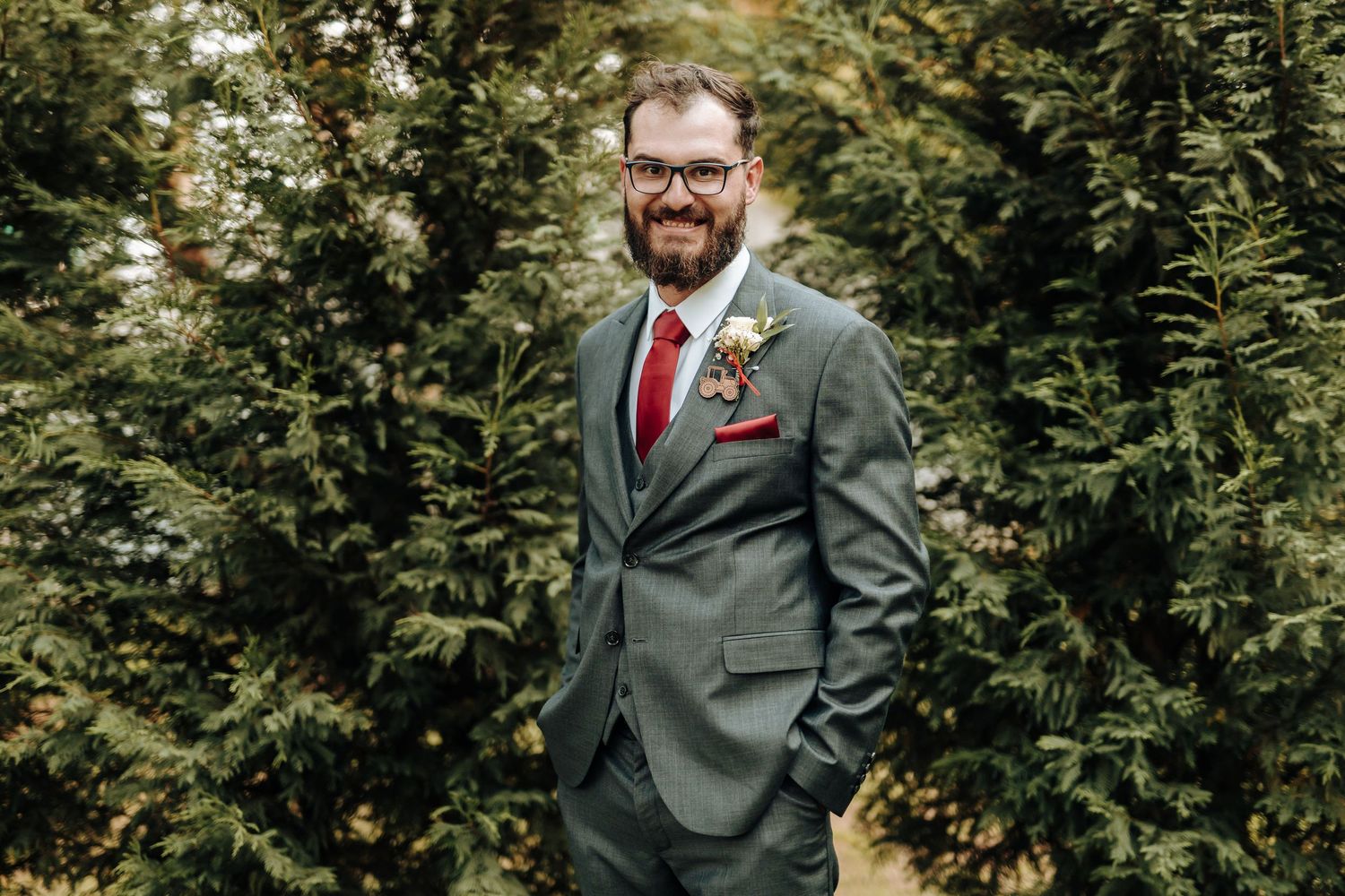 Groom in grey suit and red tie poses against green foliage backdrop.