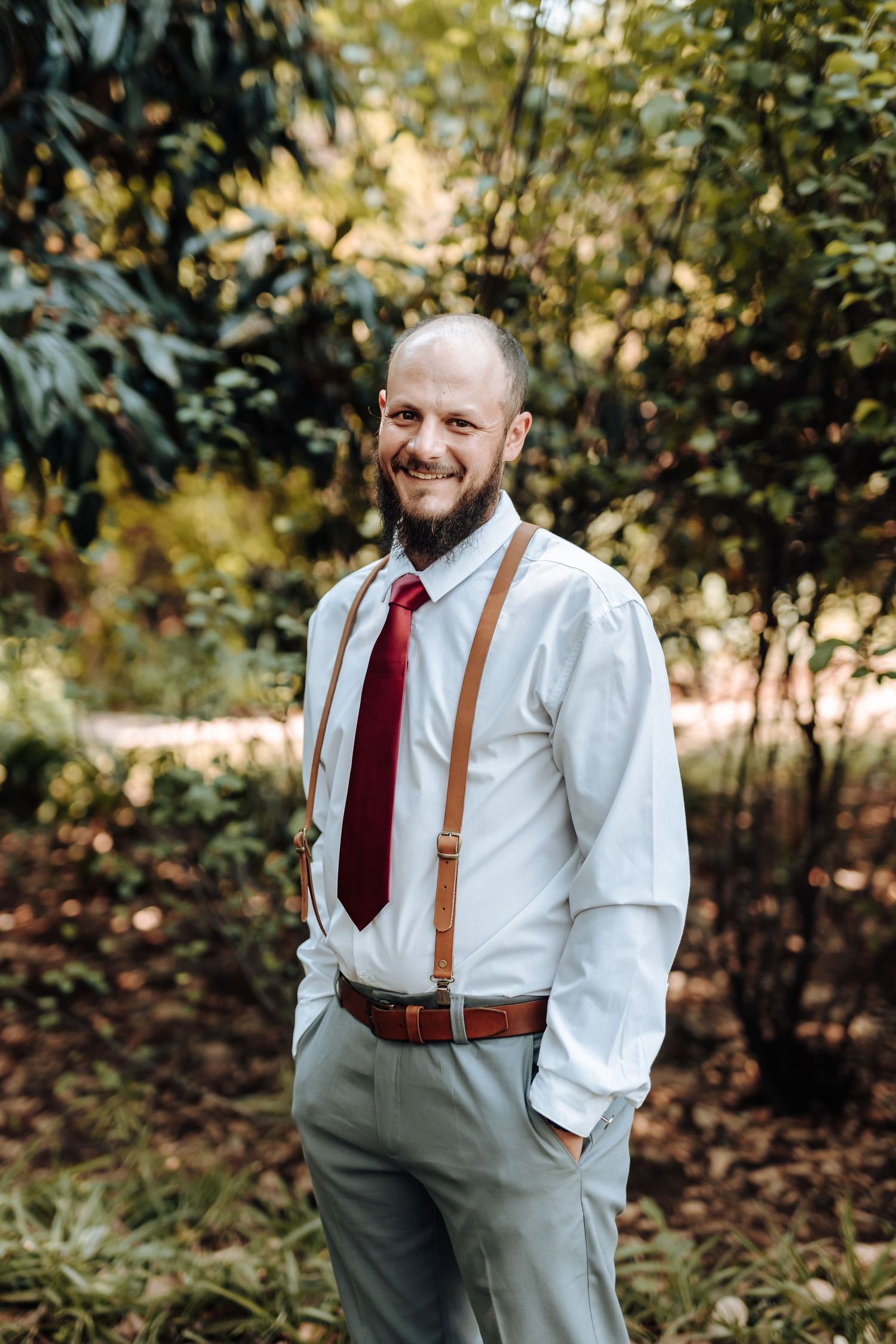 Groomsman in white shirt and suspenders smiles in outdoor garden setting.
