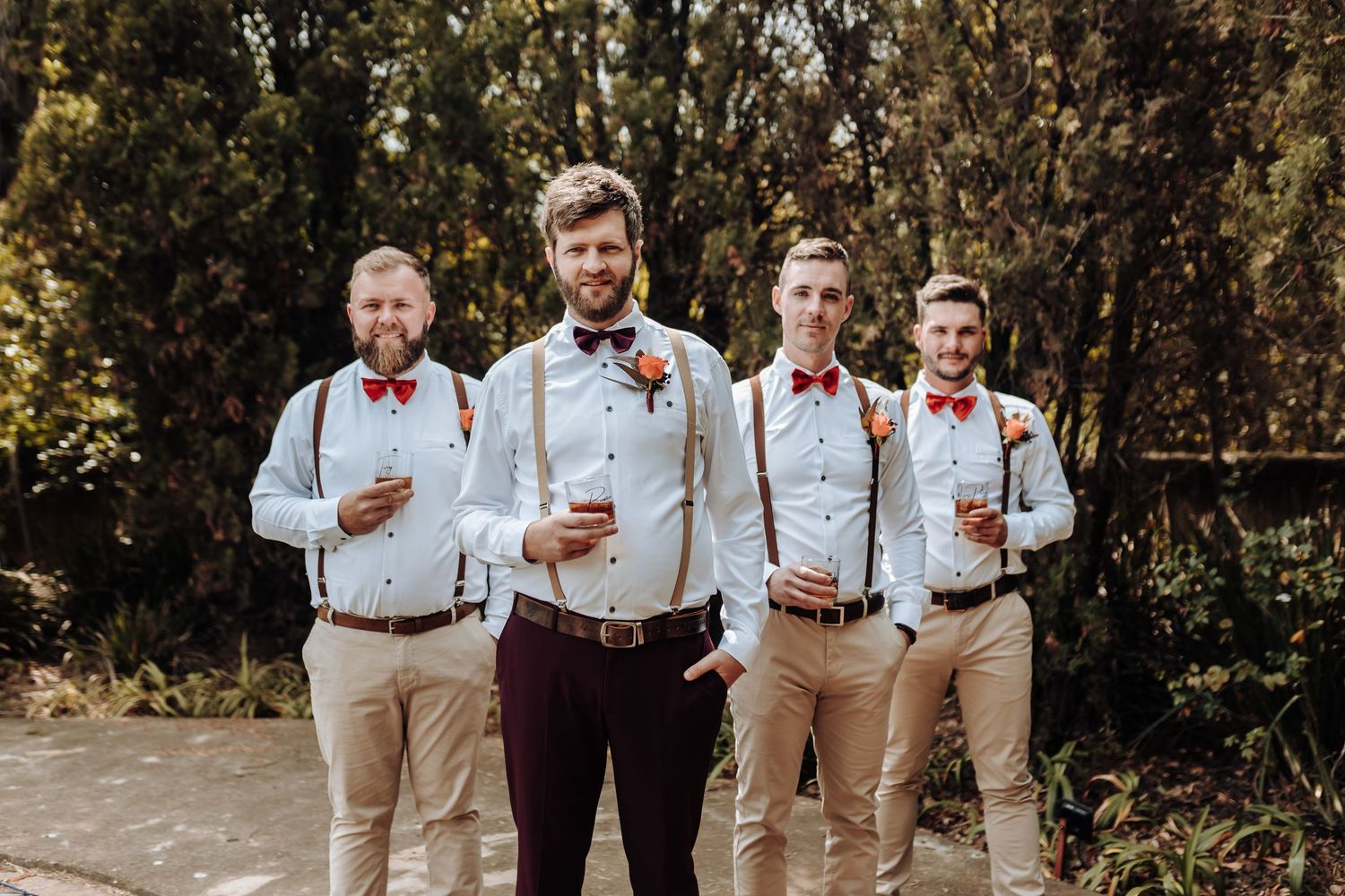 Groomsmen in suspenders and red bow ties pose with drinks outdoors.