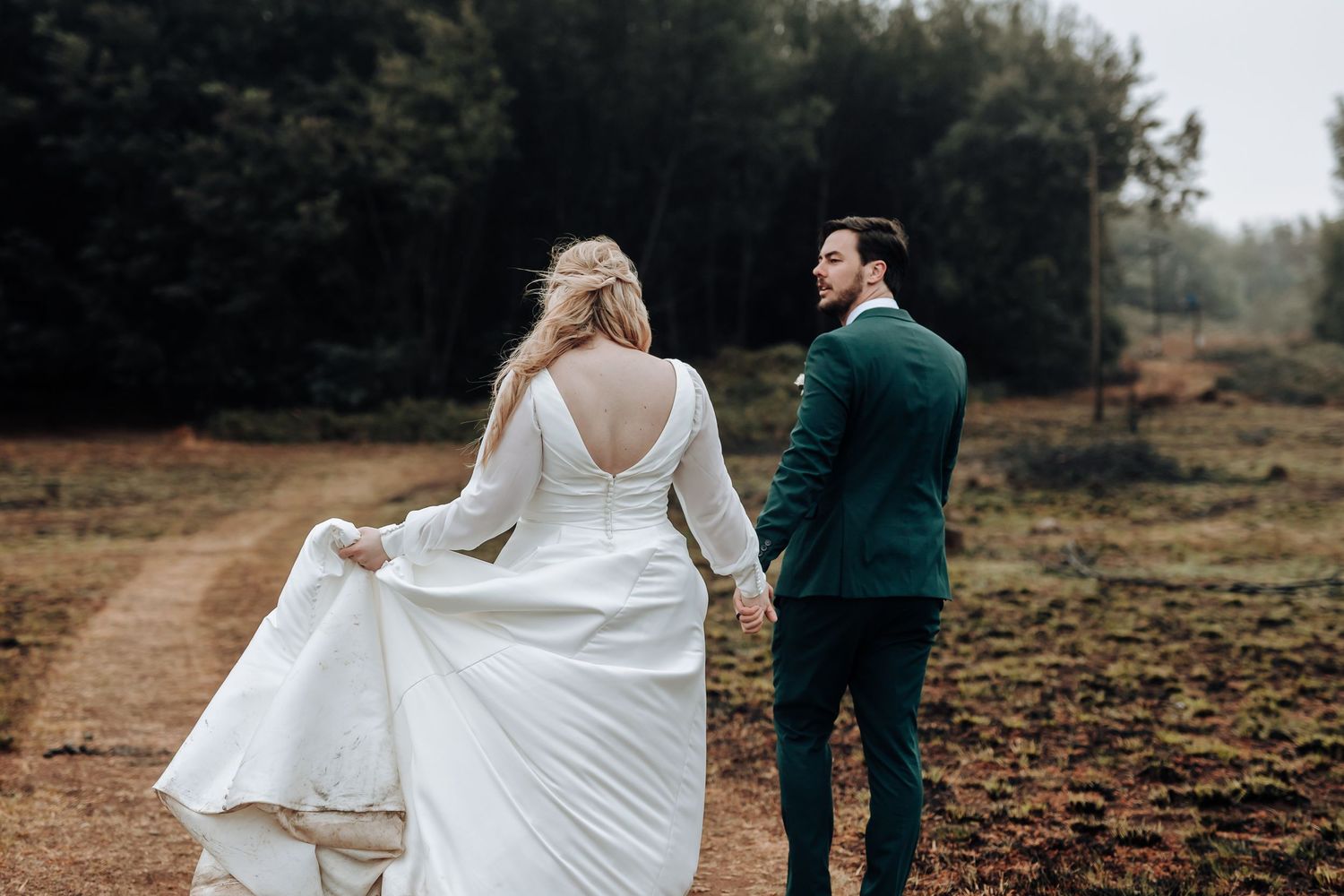 A couple in wedding attire walks hand in hand down a nature path surrounded by trees.
