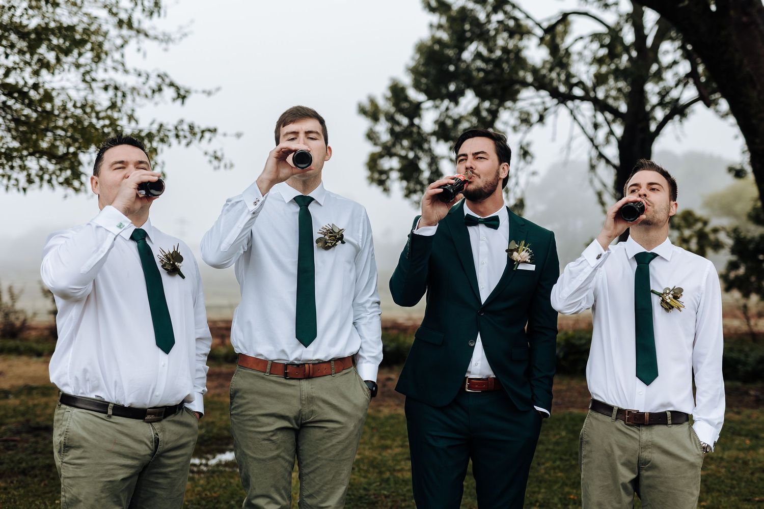 Groomsmen share a celebratory drink together while wearing matching green ties and white shirts outdoors.