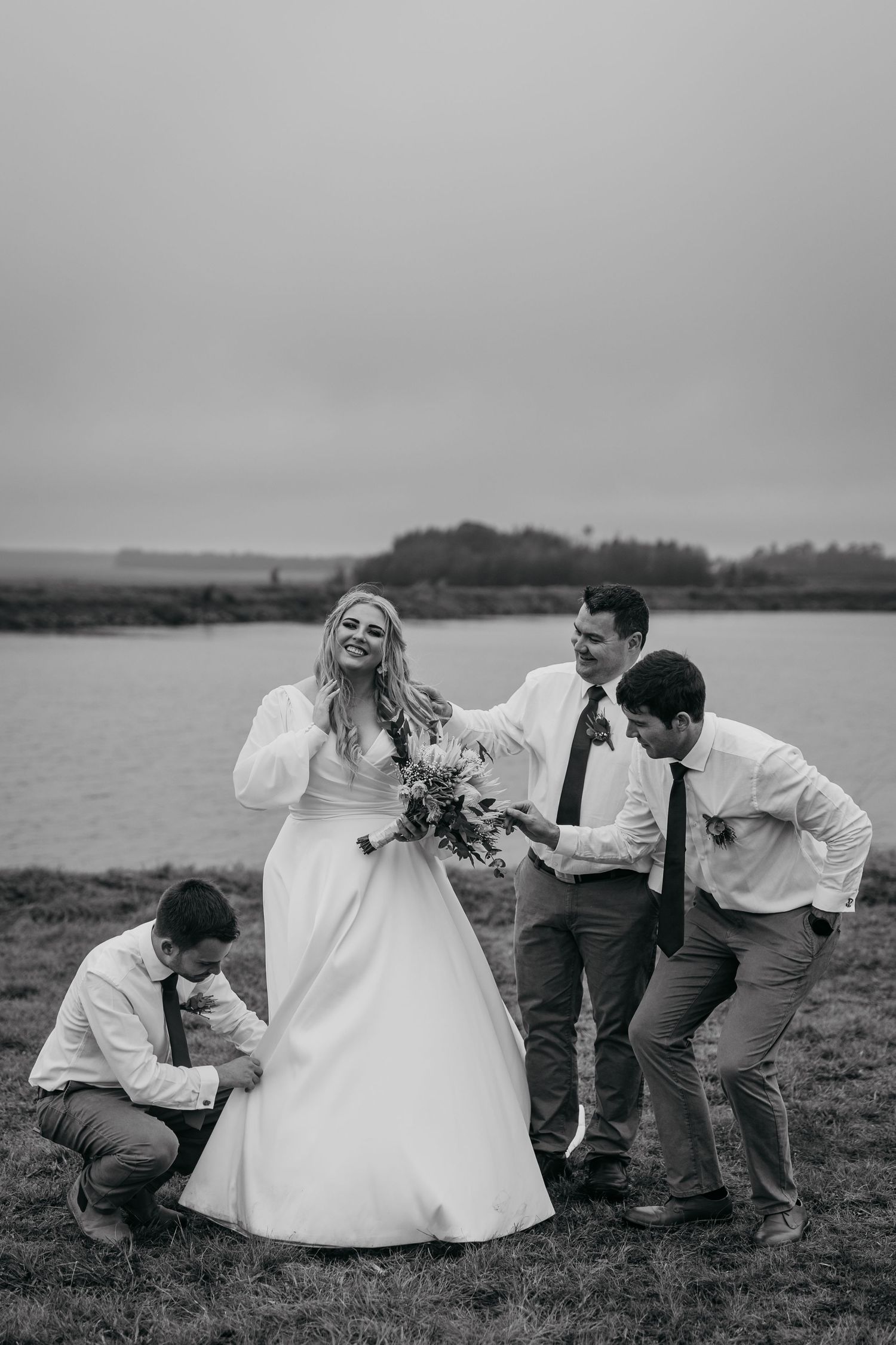 Wedding party shares a candid laughing moment by a lake in black and white photography.